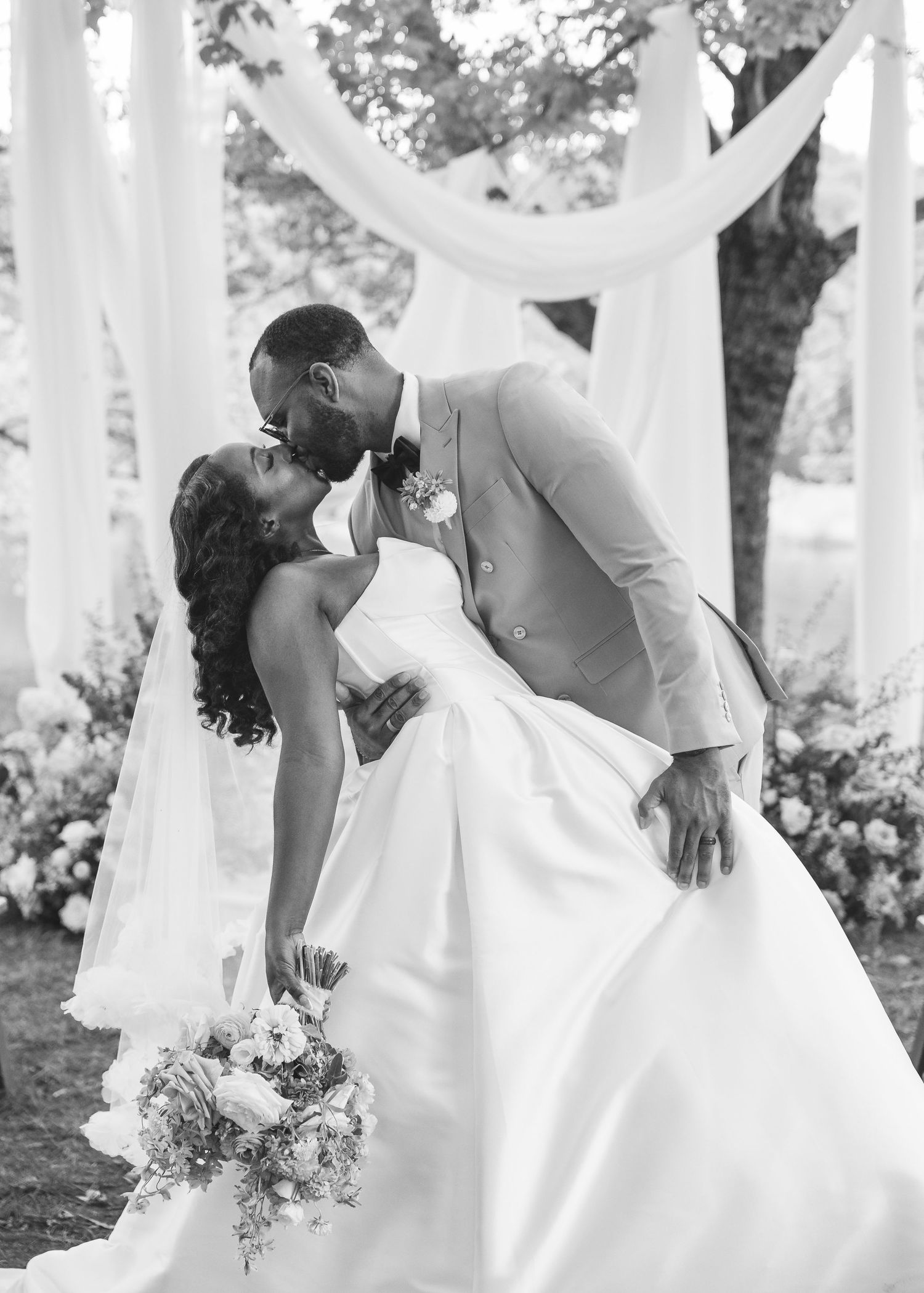 Black and white photo of bride and groom sharing a romantic dip kiss at outdoor wedding ceremony.