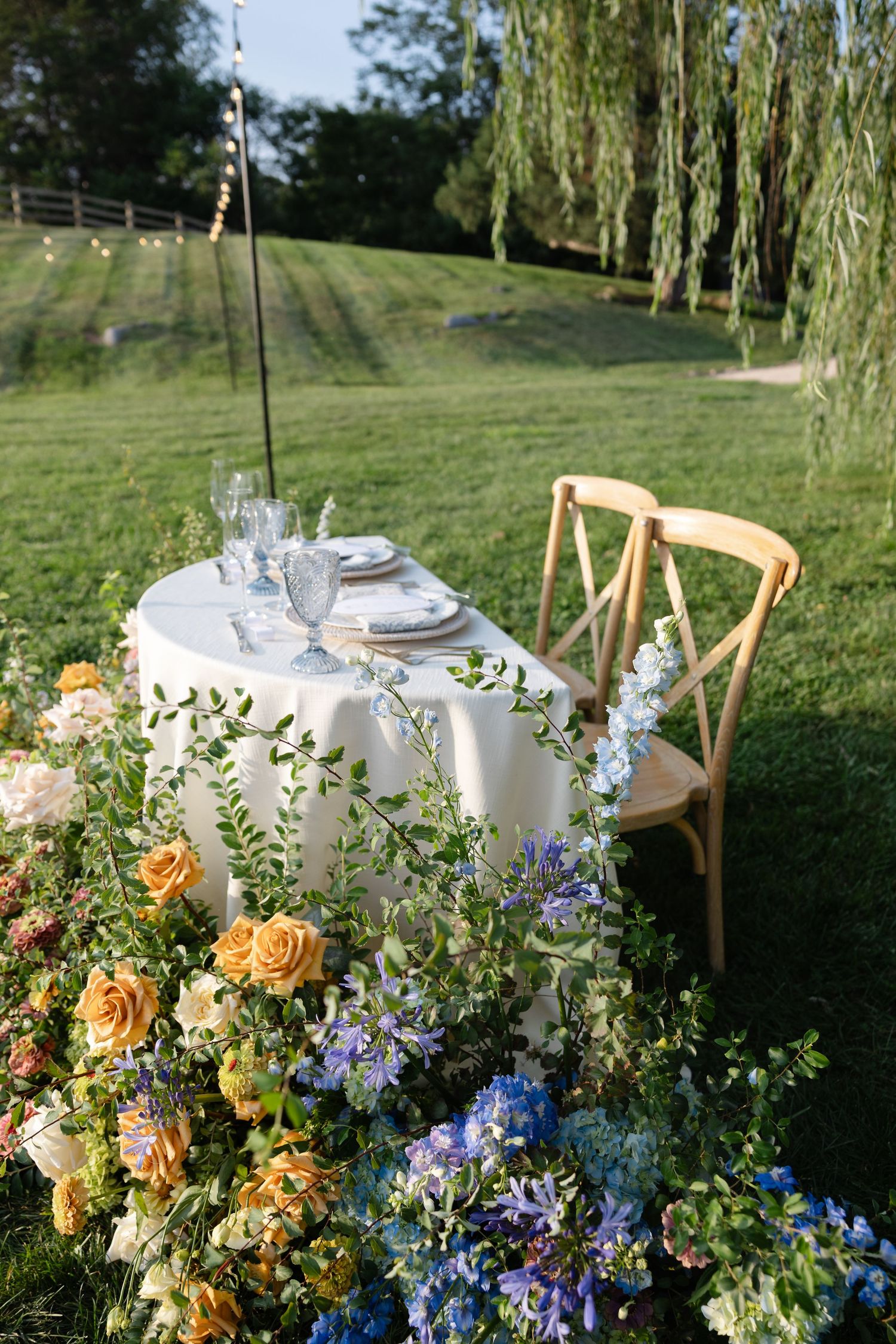 Outdoor wedding sweetheart table surrounded by lush garden flowers in yellow, blue, and white near willow trees.