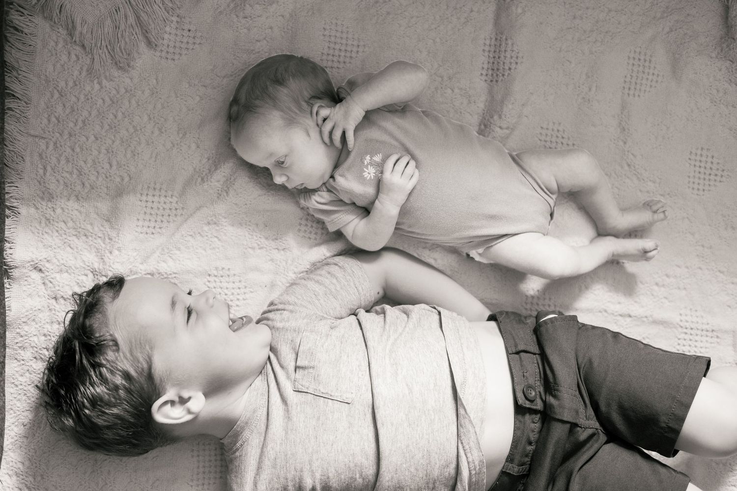 Black and white photo of young boy lying beside newborn sibling on a soft blanket.