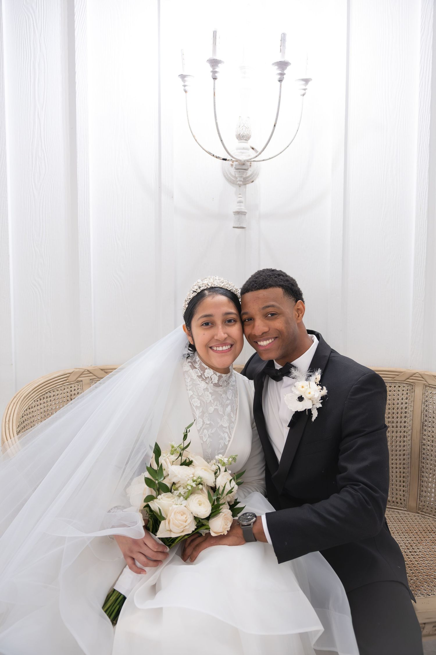 Happy newlywed couple posing on wicker bench, bride in lace veil holding white rose bouquet.