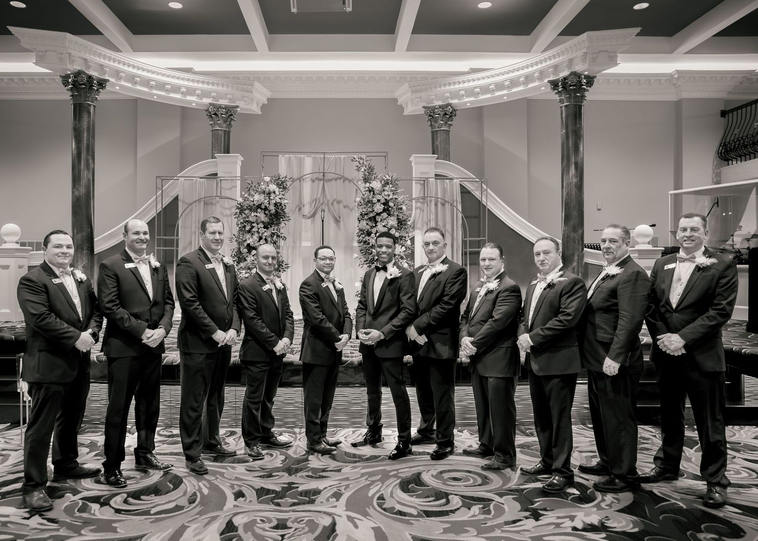 Black and white photo of groomsmen in tuxedos posing in an elegant ballroom with decorative columns.