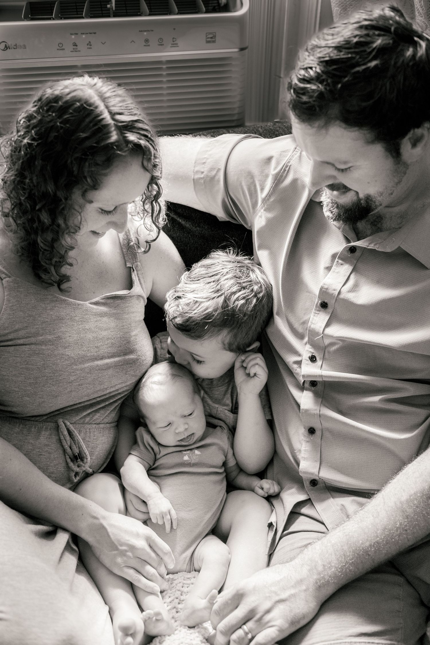 Black and white family photo of parents and toddler lovingly gazing at newborn baby together.