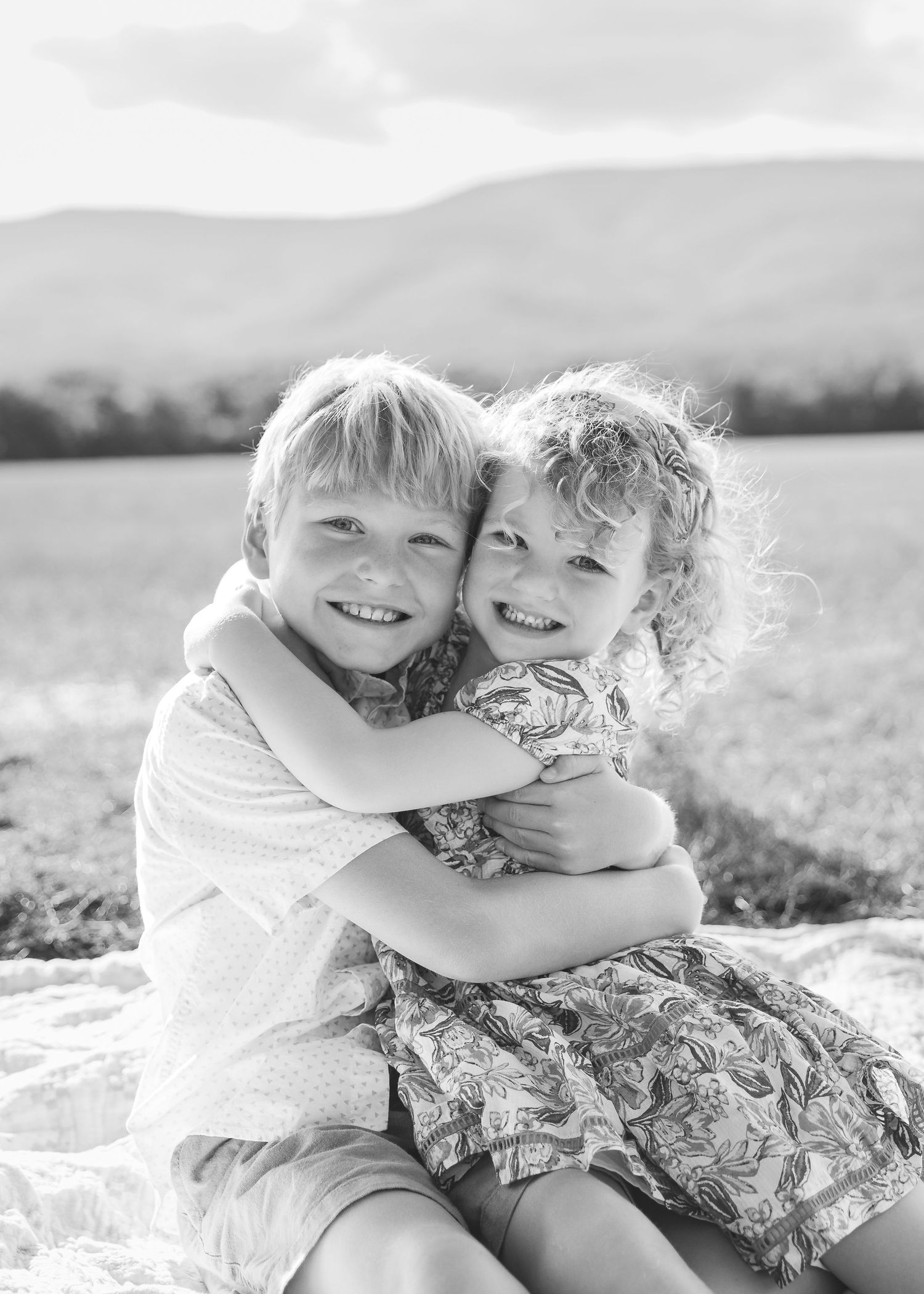 Two young siblings hugging and smiling outdoors in a black and white portrait with mountain backdrop.