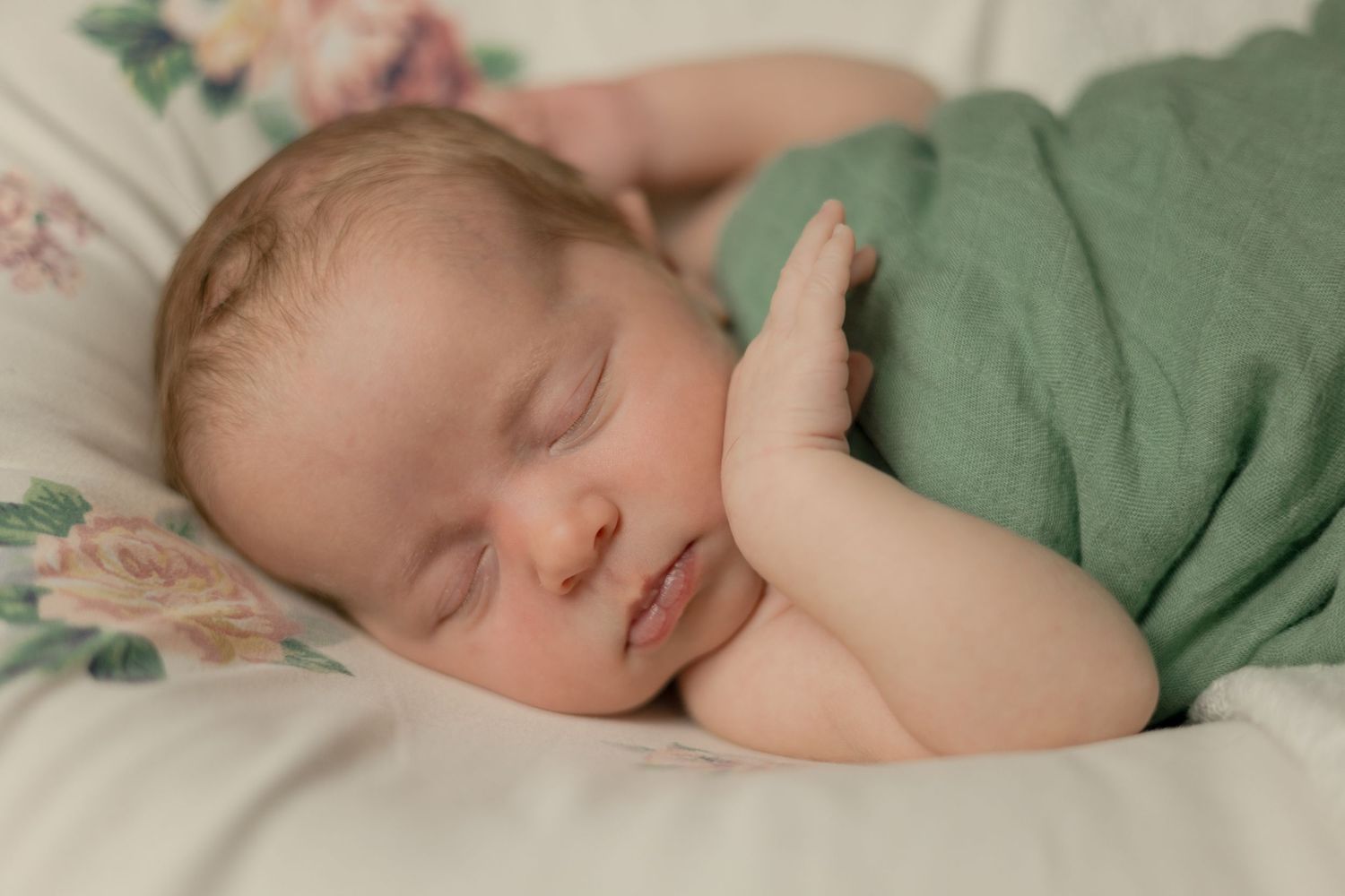 Peaceful newborn baby sleeps wrapped in soft green swaddle blanket on floral background.