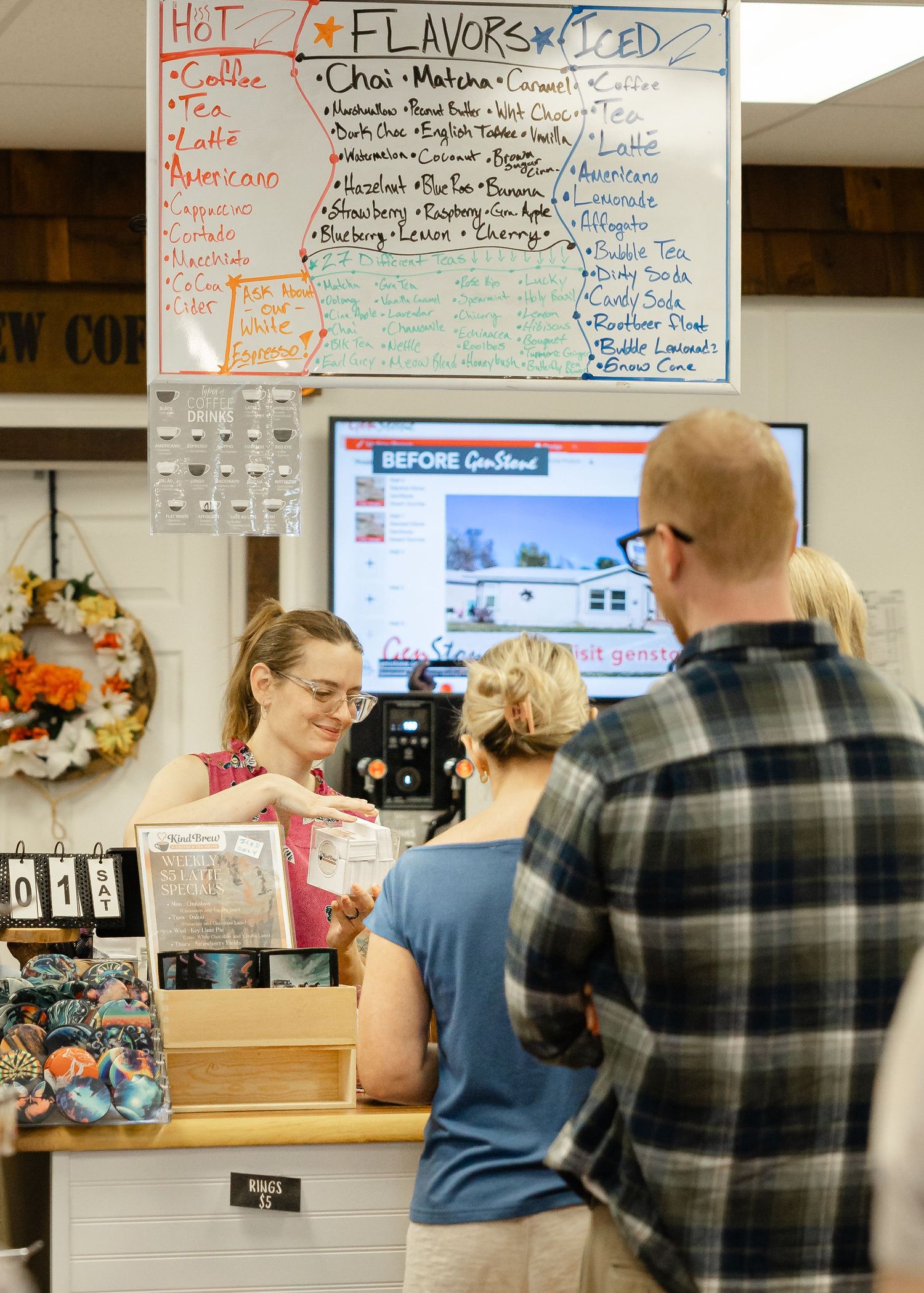 Barista serves customers at a coffee shop counter with a flavors menu board listing hot and iced drinks options.