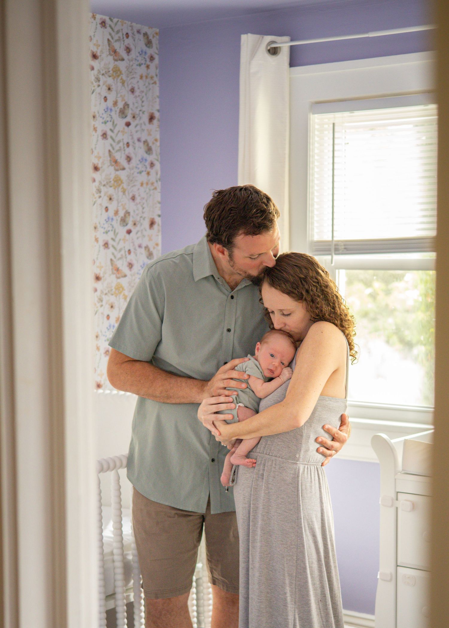 New parents tenderly holding their newborn baby in a softly lit nursery with floral wallpaper.