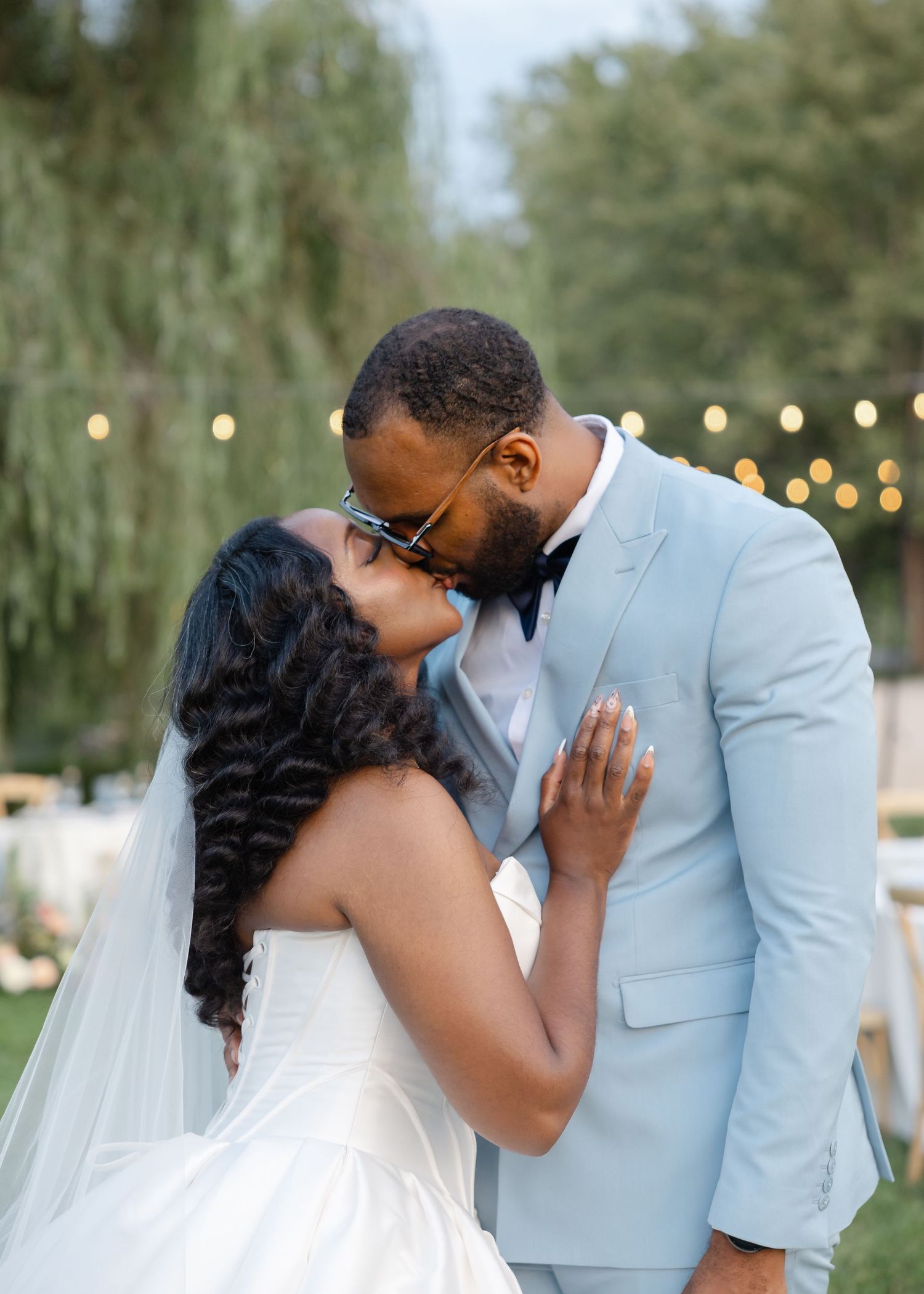Black bride and groom sharing a kiss outdoors, groom wearing light blue suit with bow tie.