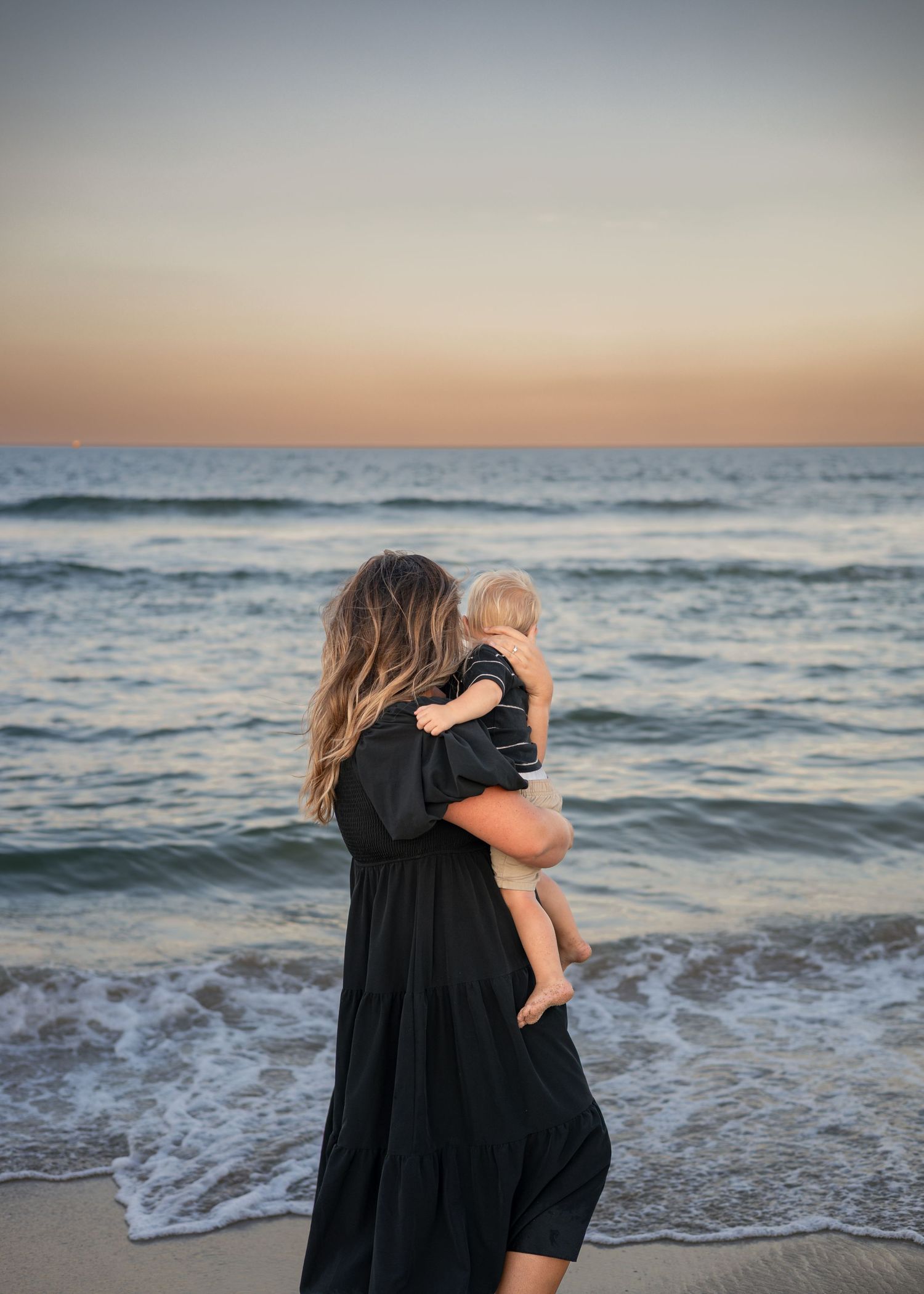 A mother in a black dress holds her baby on a beach at sunset, waves gently washing ashore.