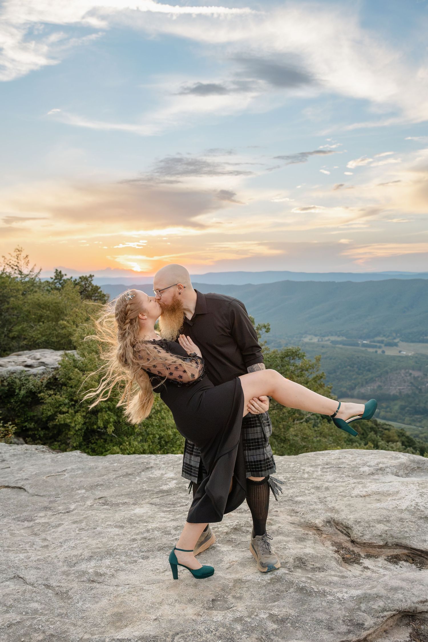 Couple sharing a romantic kiss at sunset on a mountain overlook, man lifting woman in teal heels.
