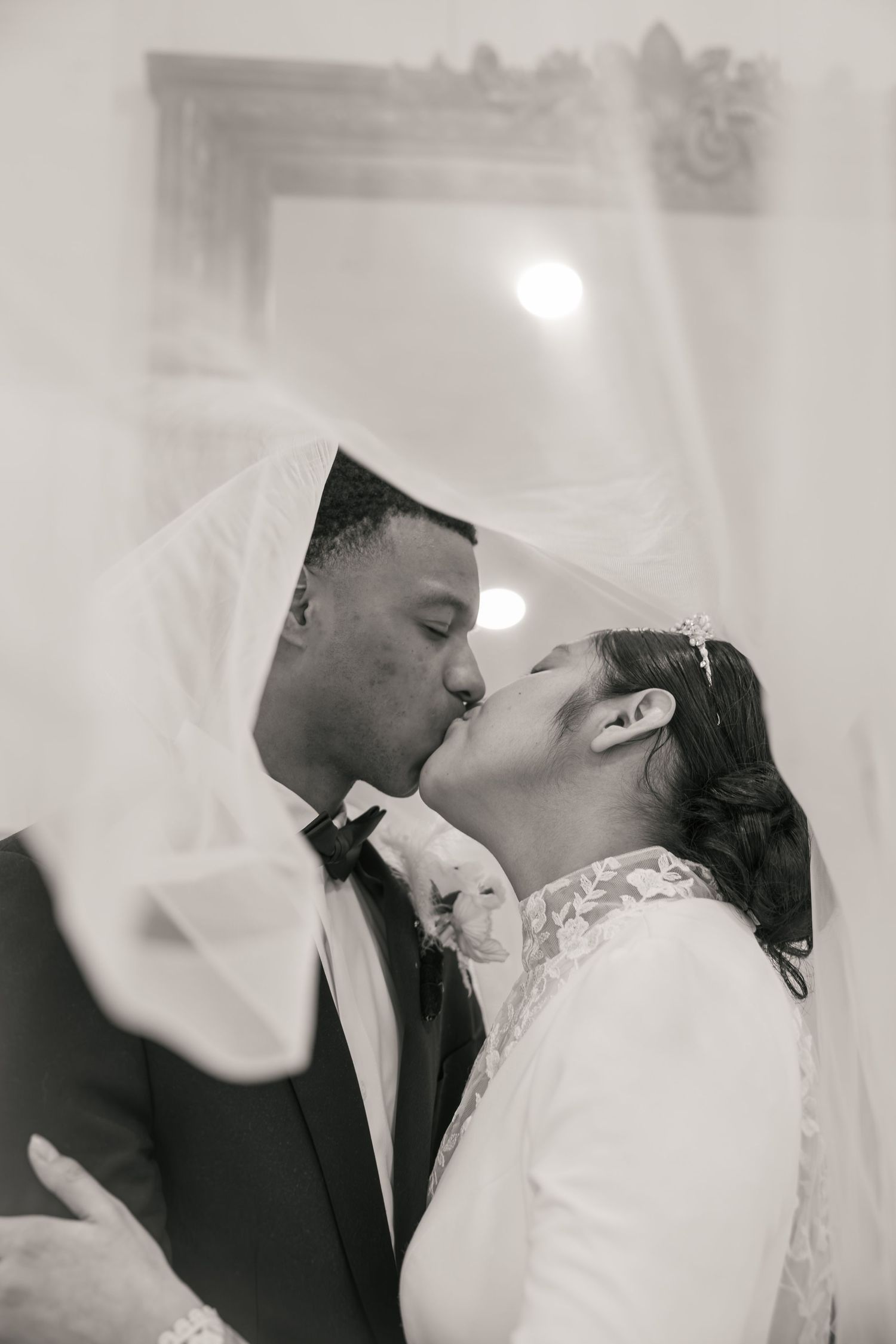 Black and white romantic photo of newlywed couple kissing under flowing bridal veil at wedding.