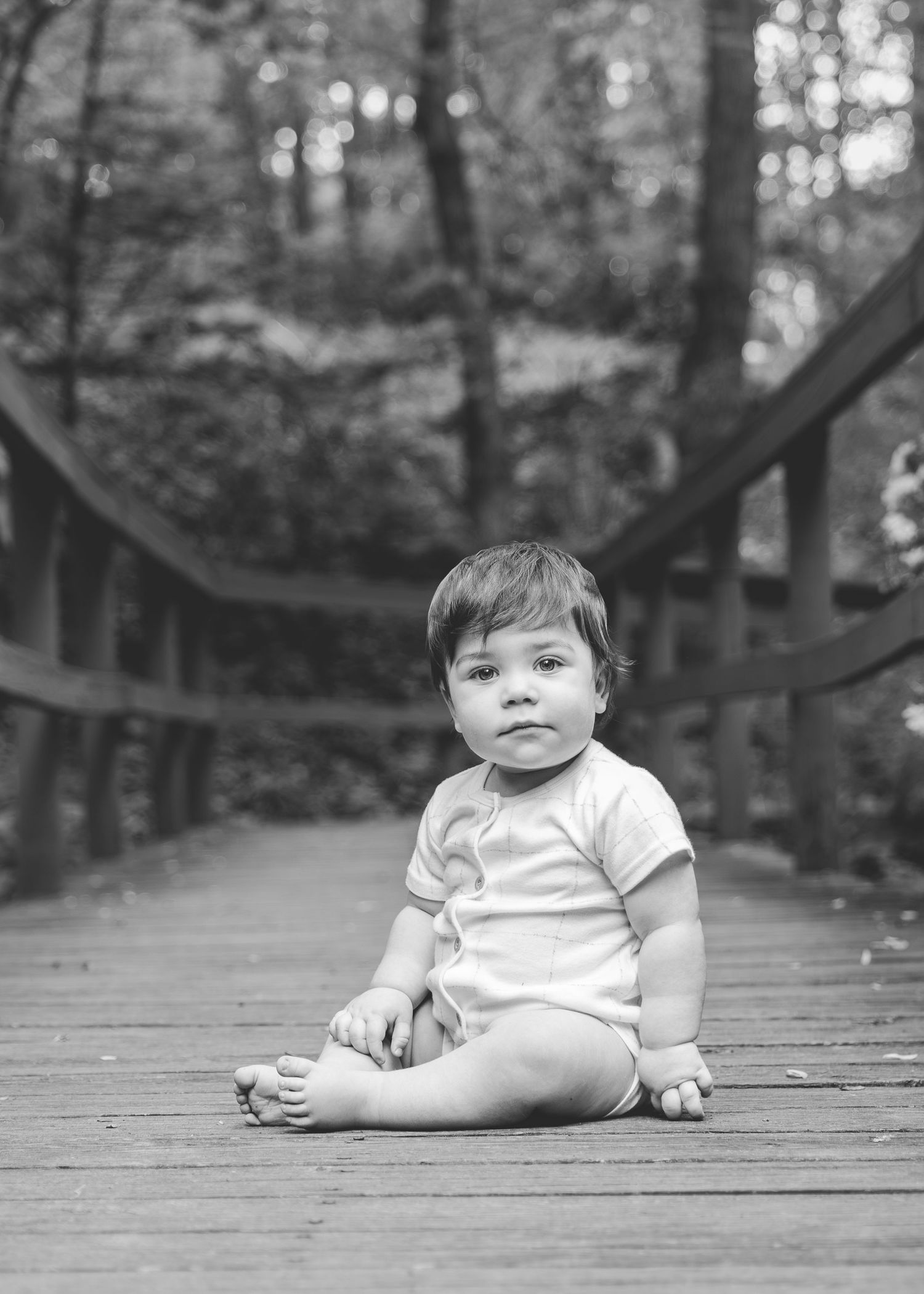 Black and white portrait of a baby sitting on a wooden bridge surrounded by trees.