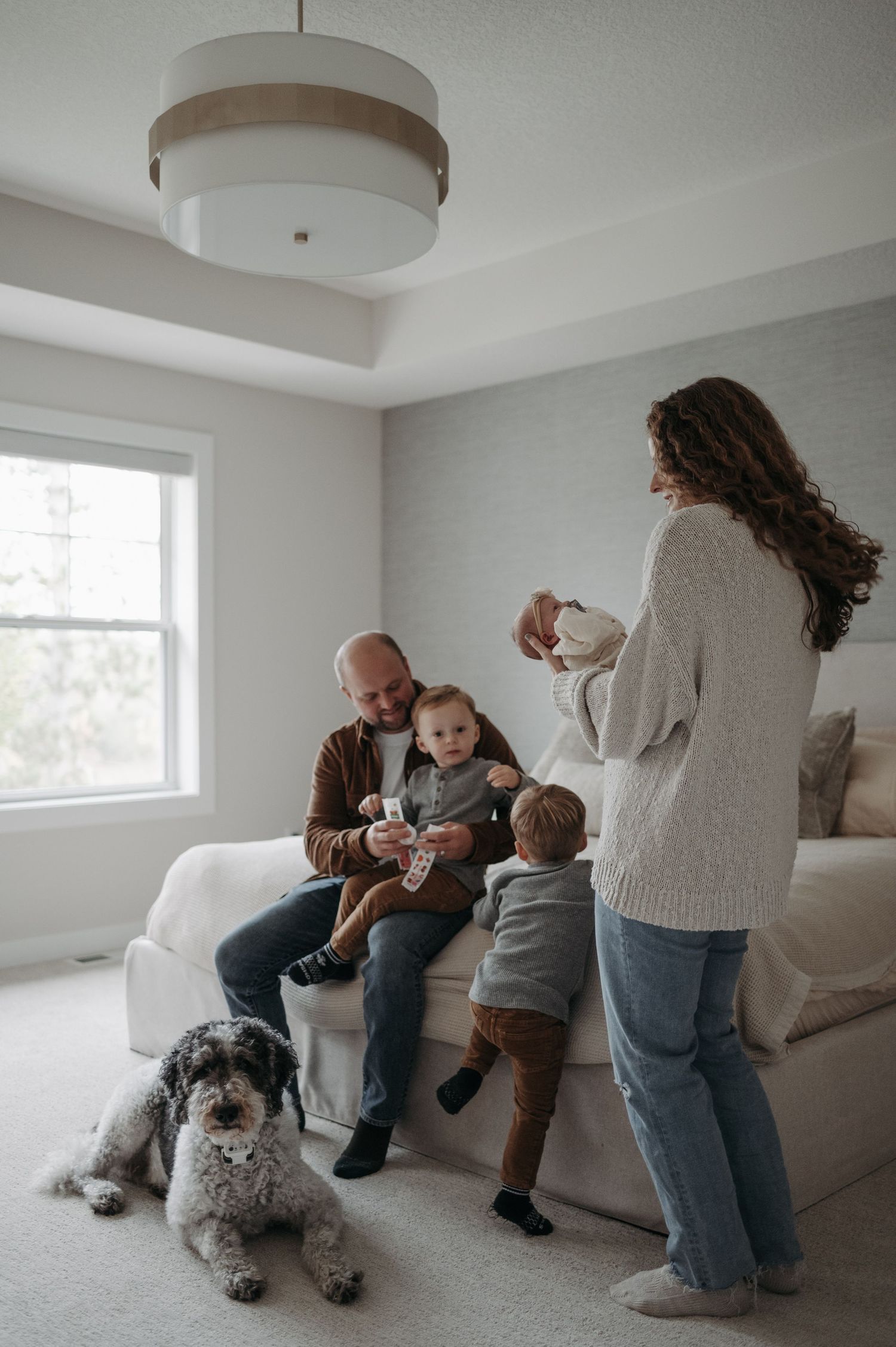 Family with two toddlers and a fluffy dog enjoying a playful moment in a bright, modern bedroom.