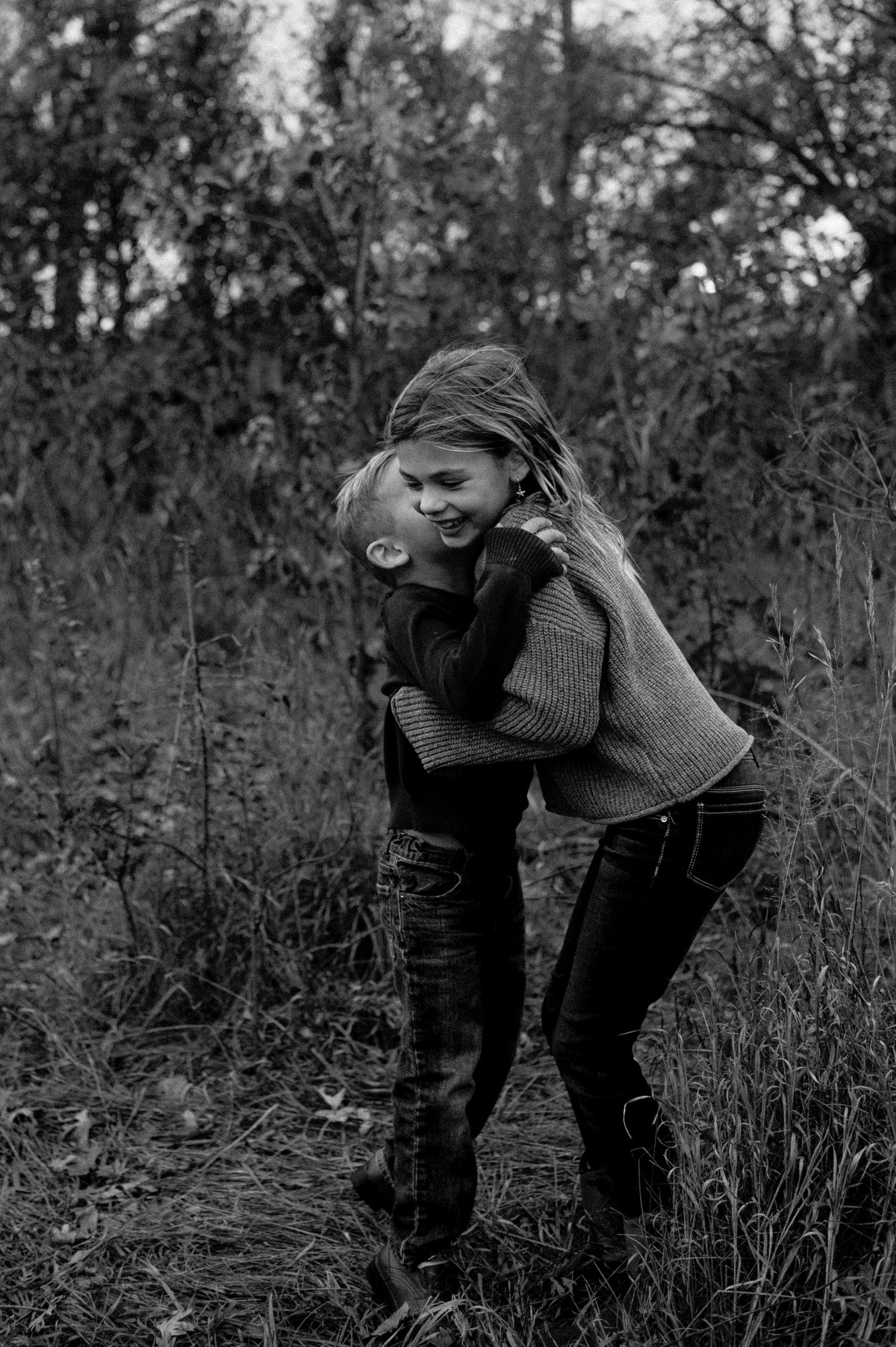 A boy hugs and kisses a laughing girl in a black and white outdoor portrait among wild grass. Stillwater, Minnesota.