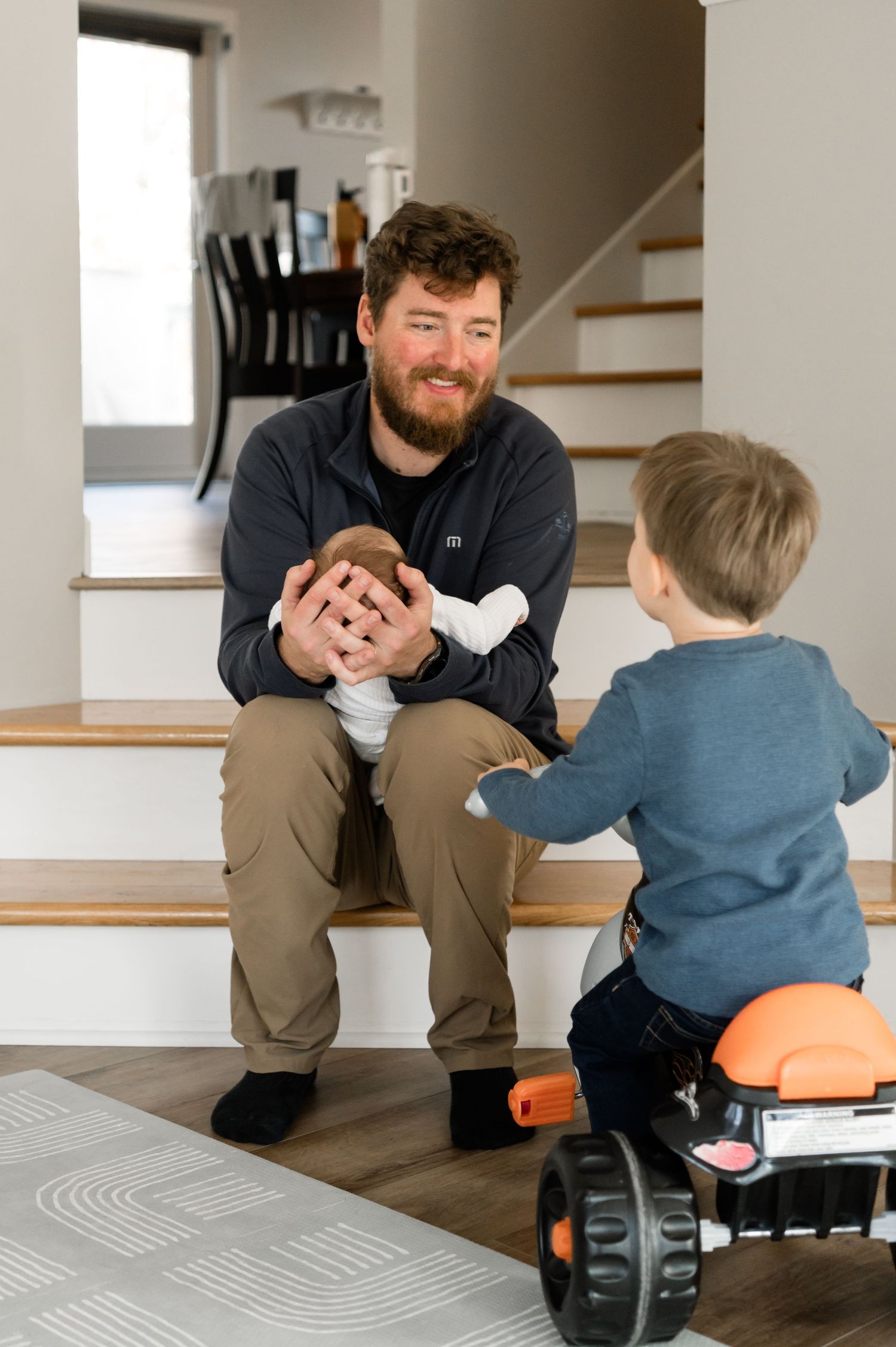 Bearded father holding newborn sits on stairs smiling at toddler on orange ride-on toy.