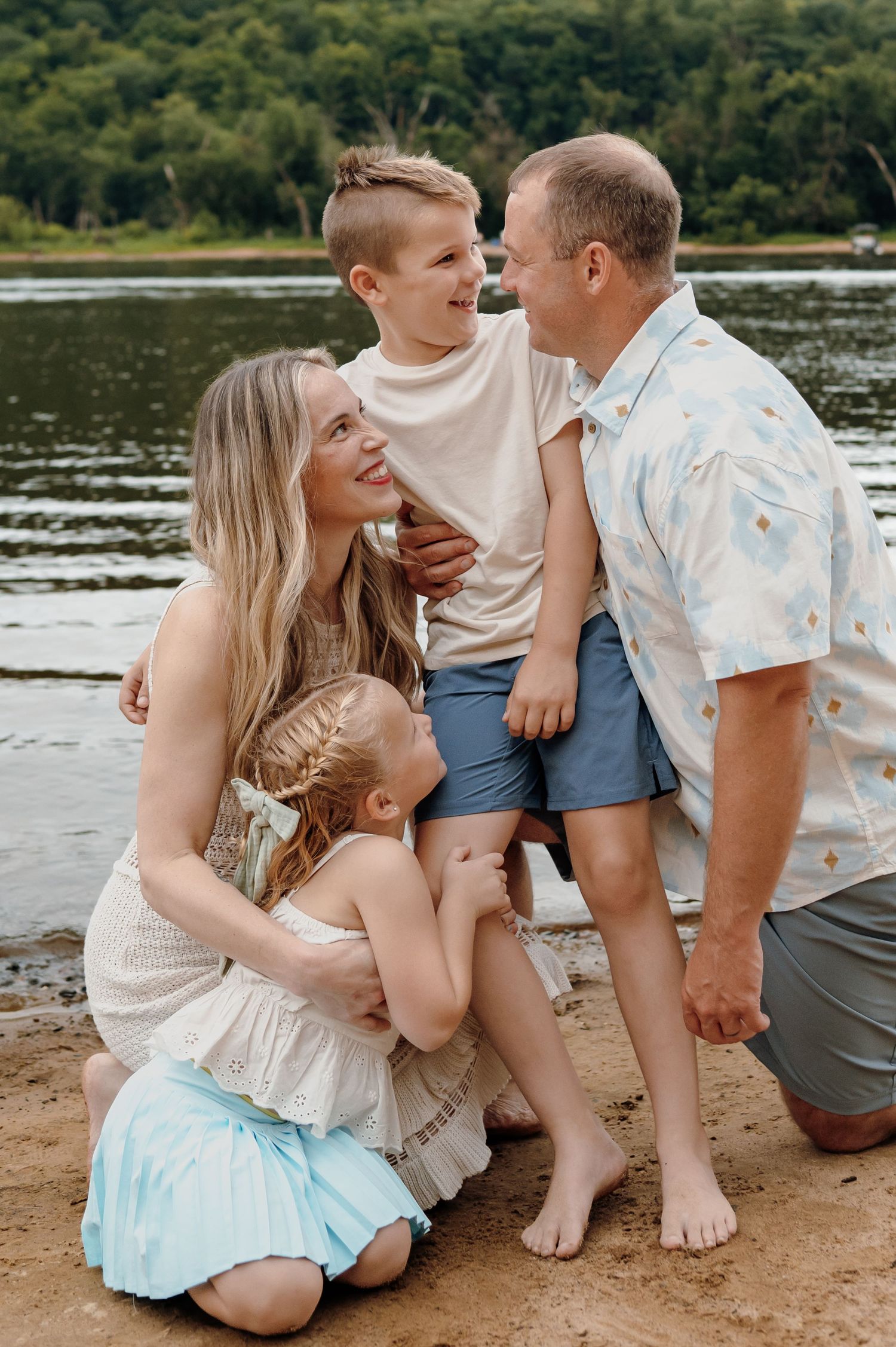 A smiling family of four poses together on a sandy riverbank.