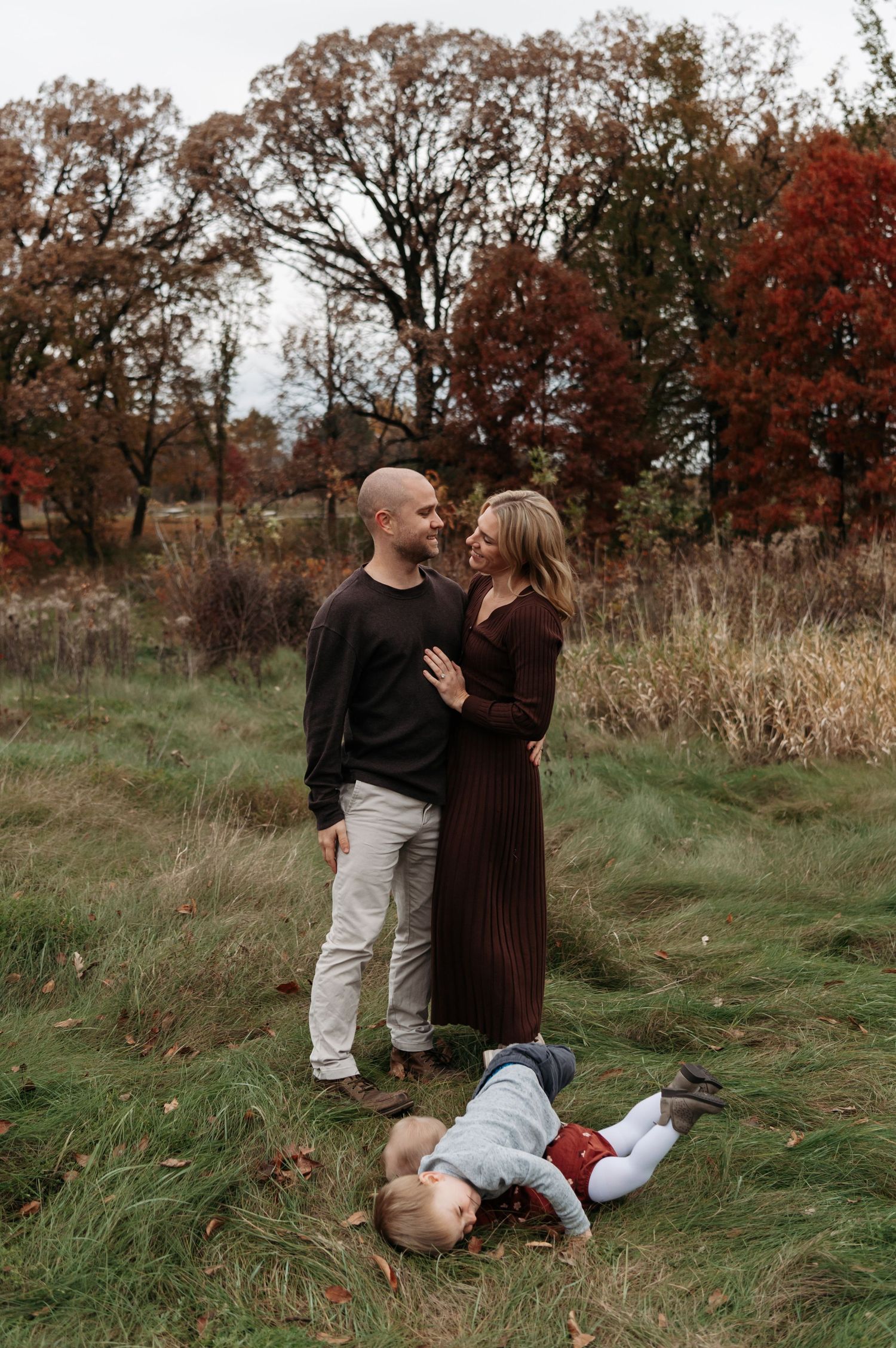 A couple shares a loving gaze in a fall meadow while their toddler plays on the ground. Lake Elmo, Minnesota.