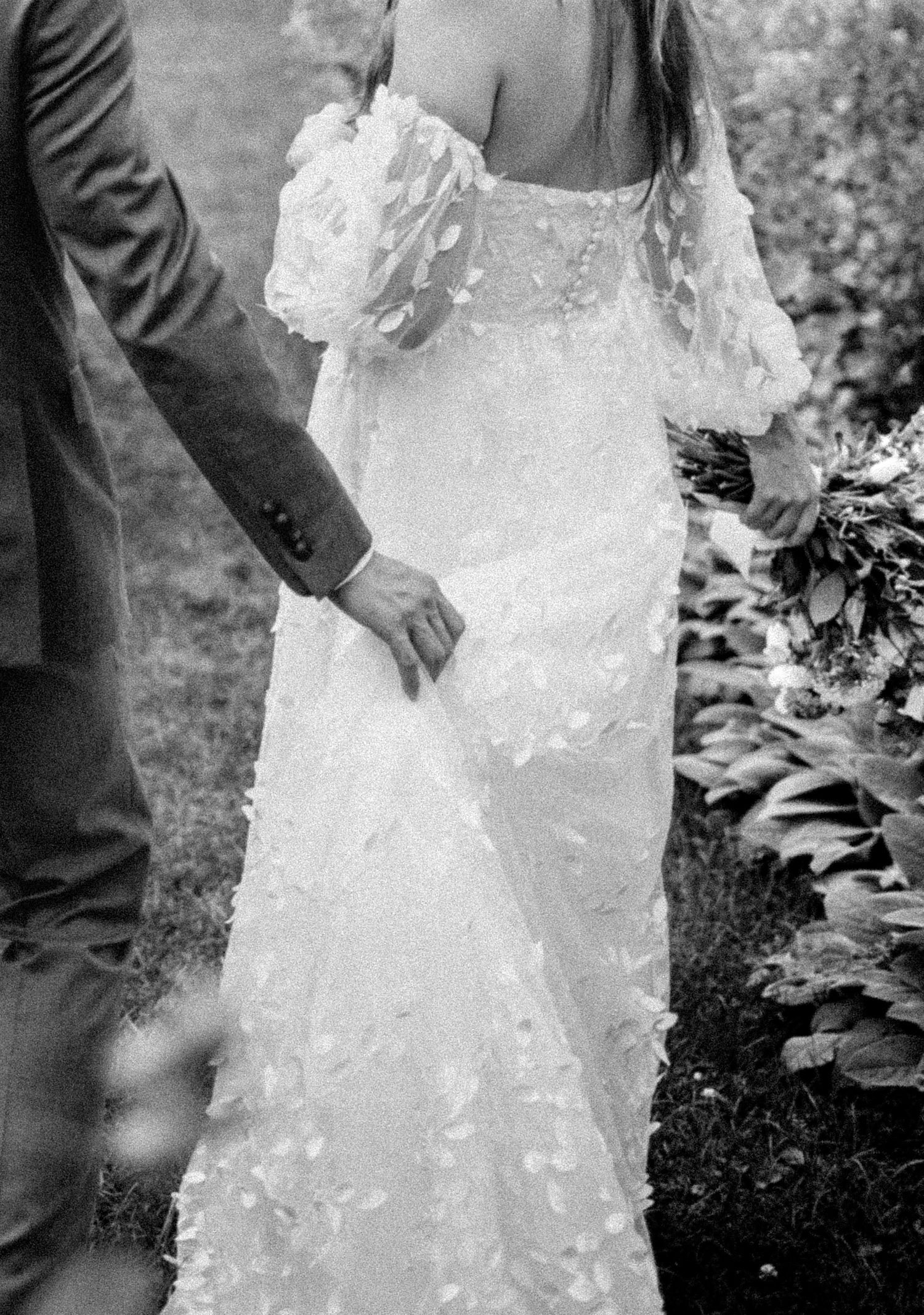 Black and white photo of bride in floral applique gown holding grooms hand in garden.