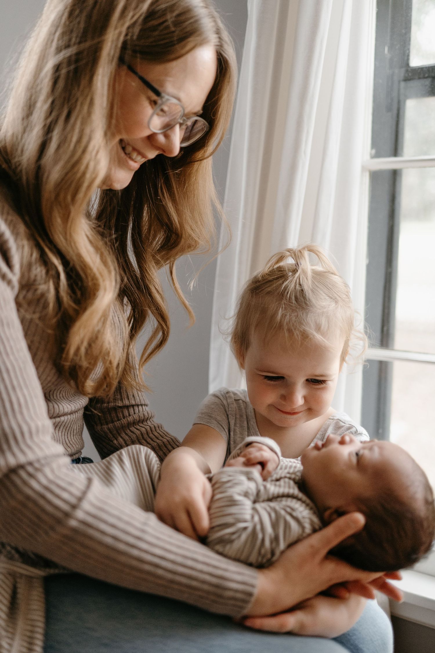 A smiling mother and toddler daughter look at a newborn baby by a window.