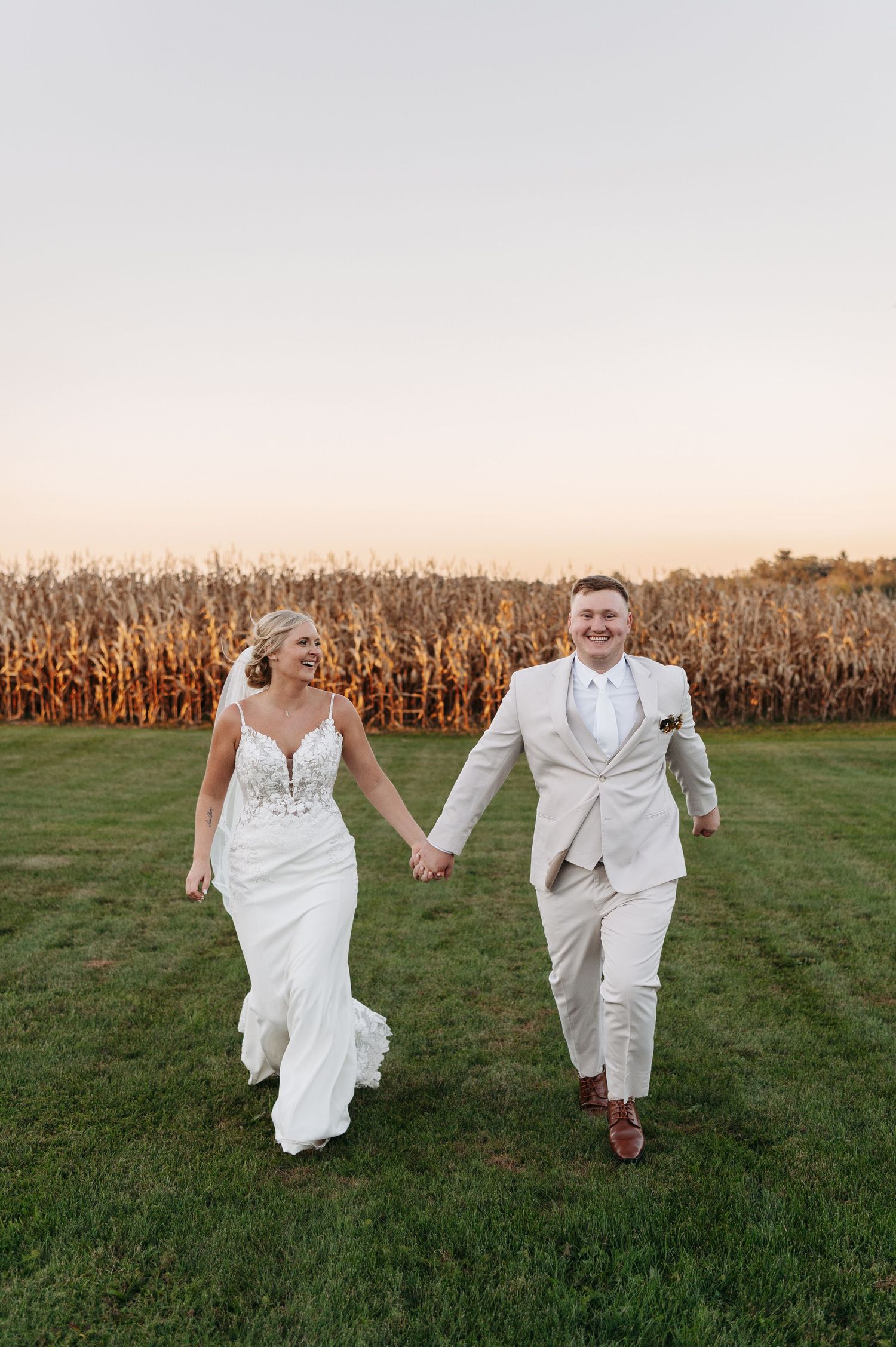 Joyful bride and groom in tan suit hold hands walking through field at golden hour farm wedding. Wisconsin. Minnesota.