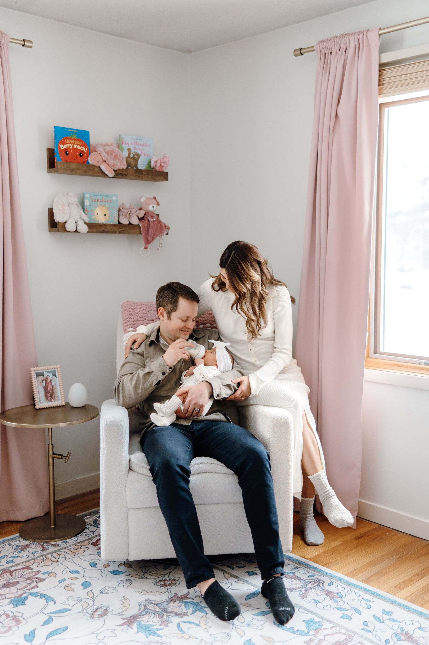 New parents admiring their newborn baby in a pink-themed nursery with a glider chair.