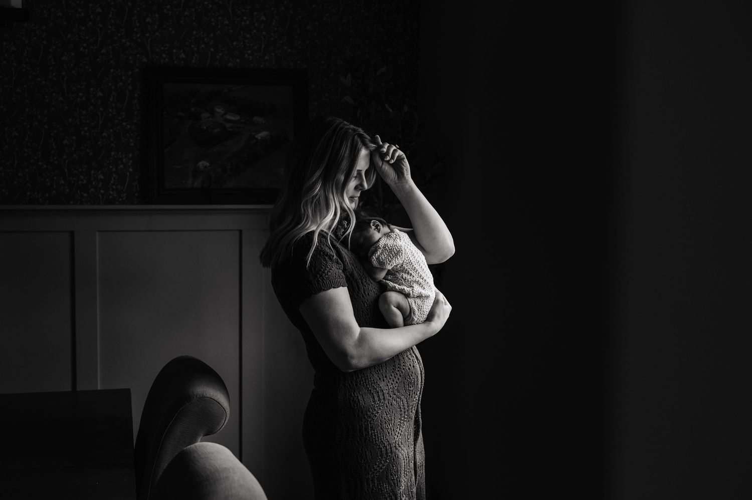 Black and white photo of mother standing and cradling newborn baby in dimly lit room.