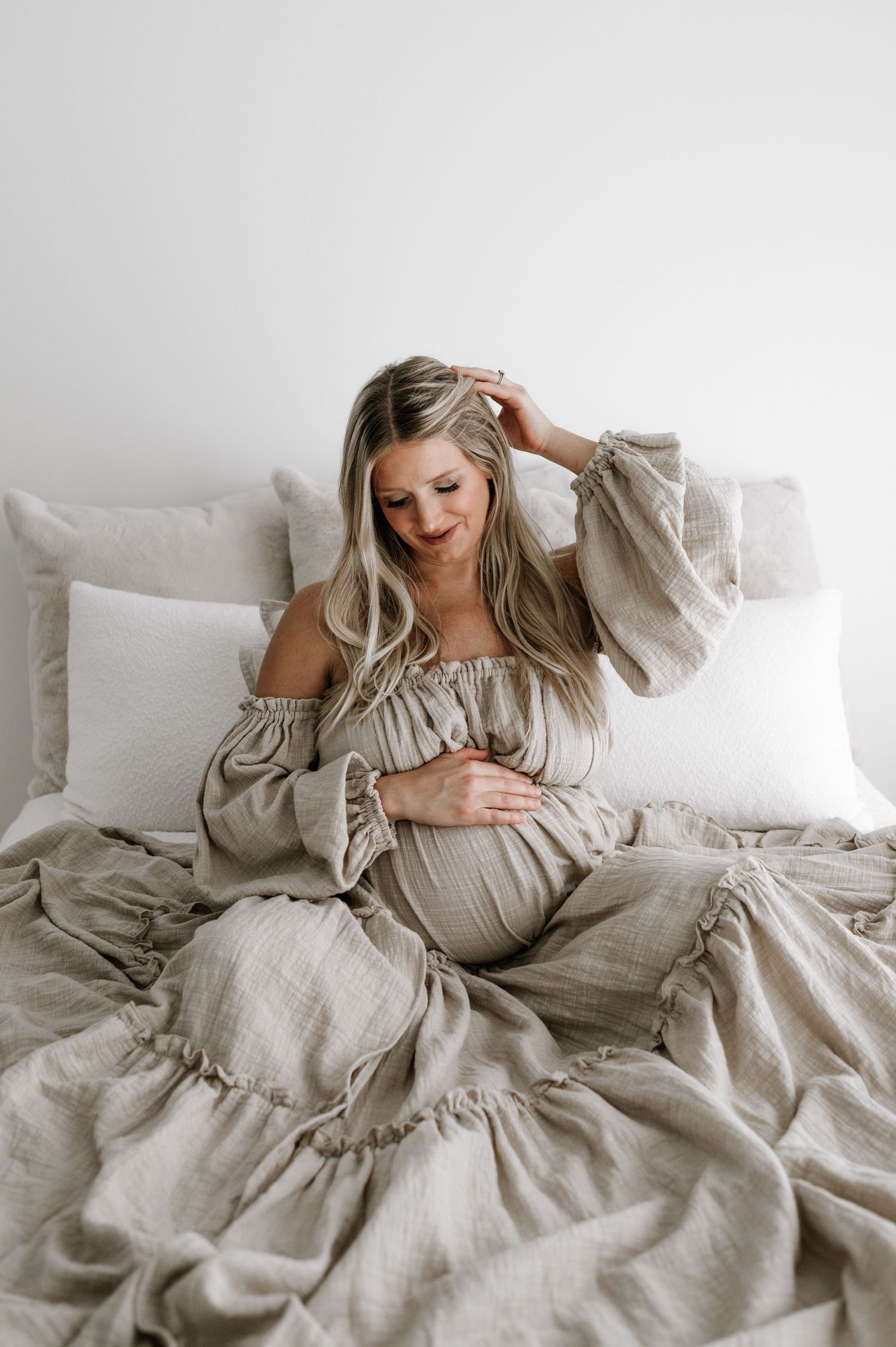 Beautiful pregnant woman in a linen off-shoulder dress sitting on a white bed. maternity photography. Wisconsin. Minnesota.