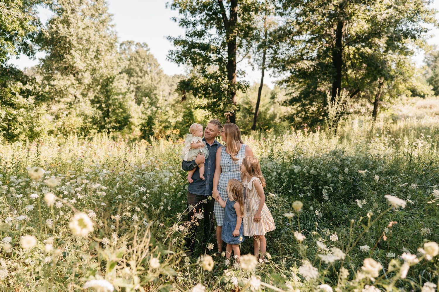 A family stands together in a sunny wildflower meadow surrounded by trees.