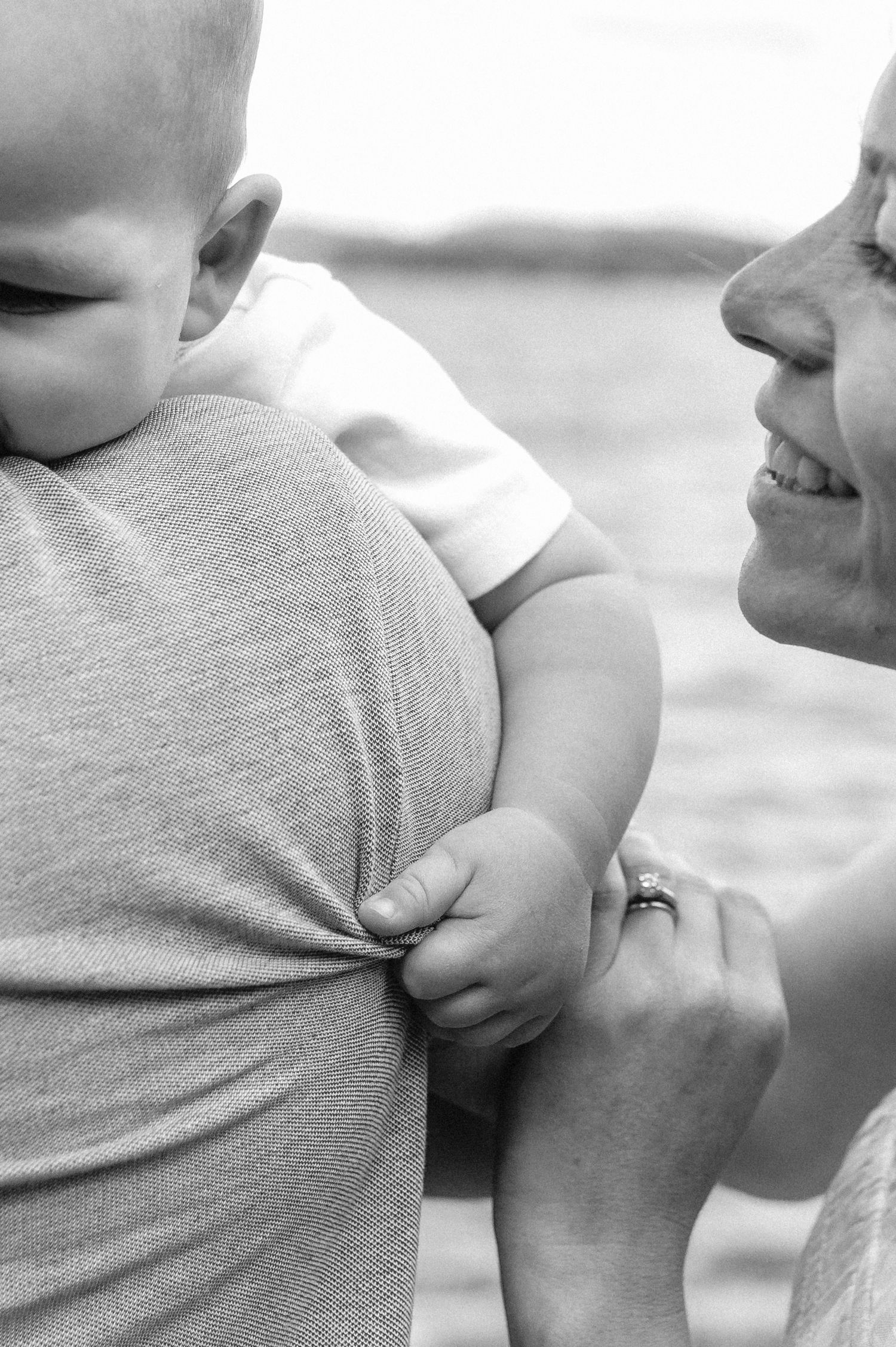 Black and white close-up of smiling mother watching father hold baby near water.