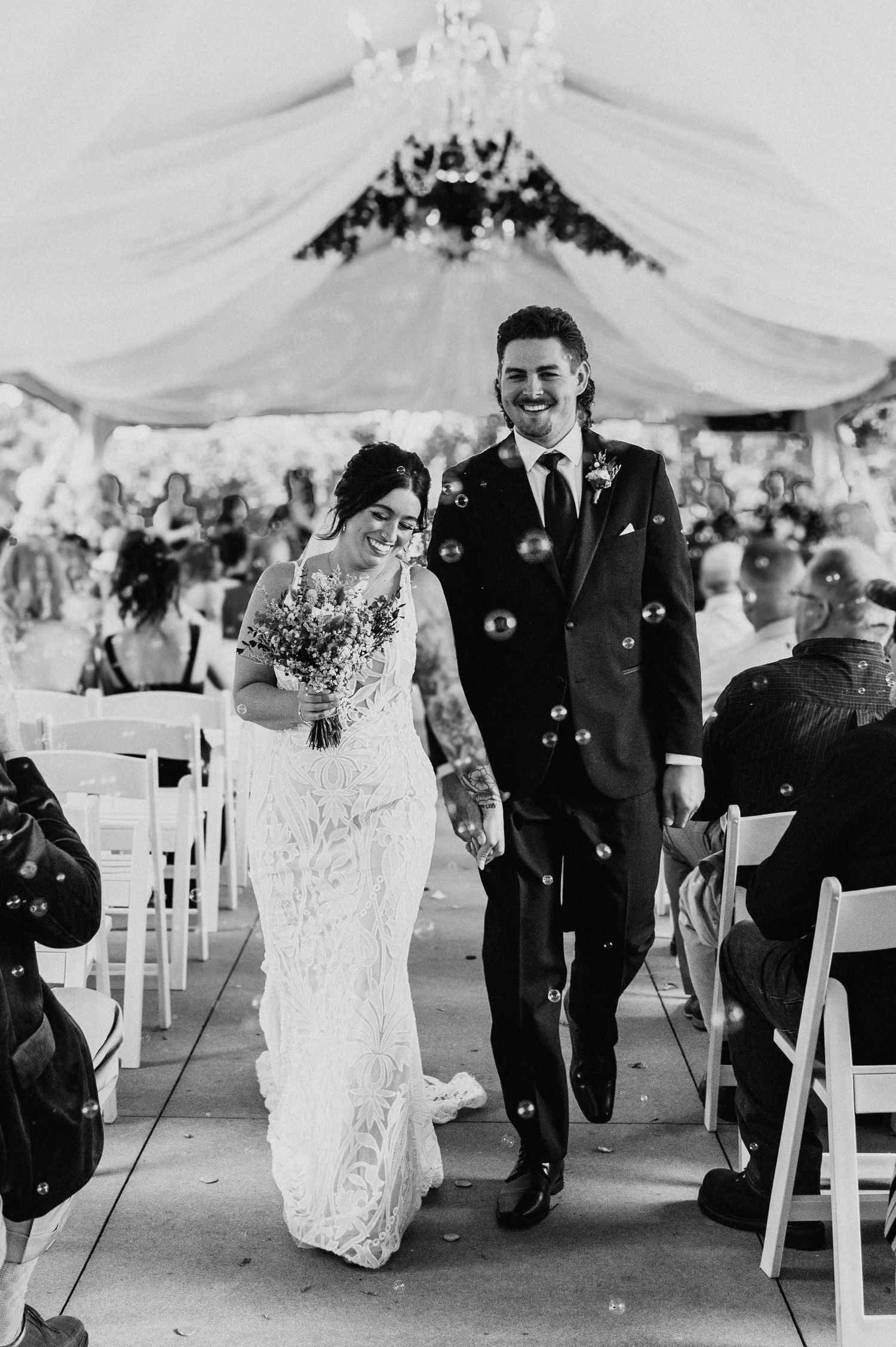 Black and white photo of joyful newlyweds walking down aisle through bubbles after ceremony.