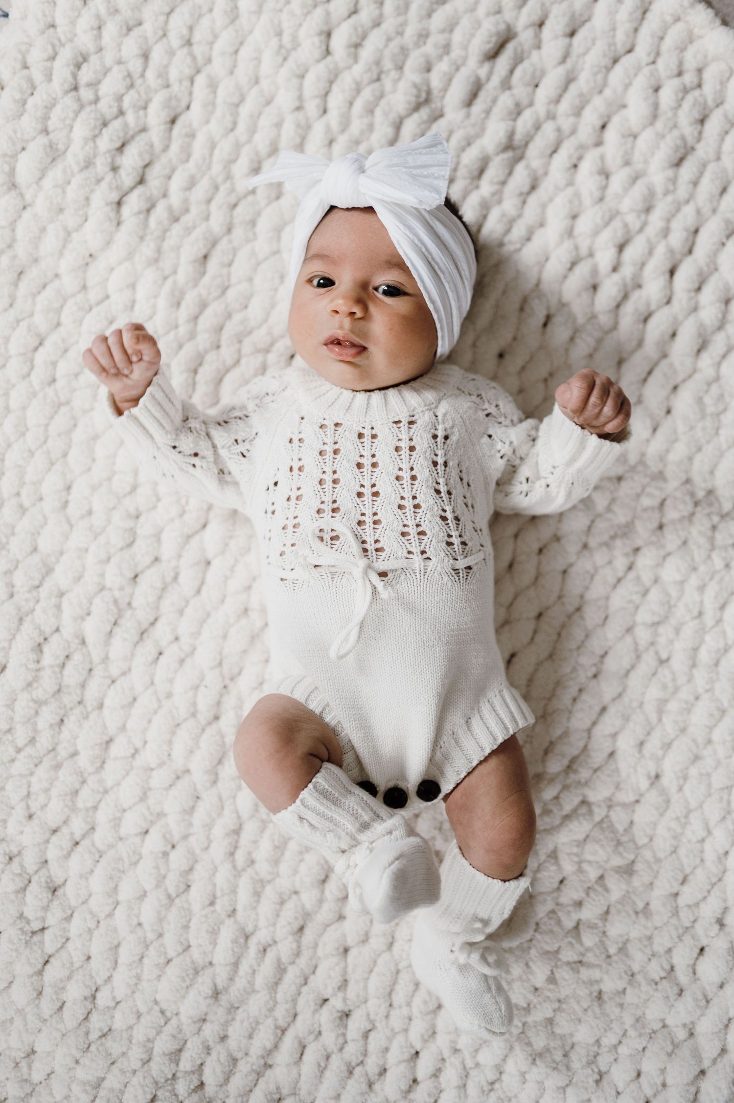 Newborn baby in white knit romper and turban headband lying on textured blanket.