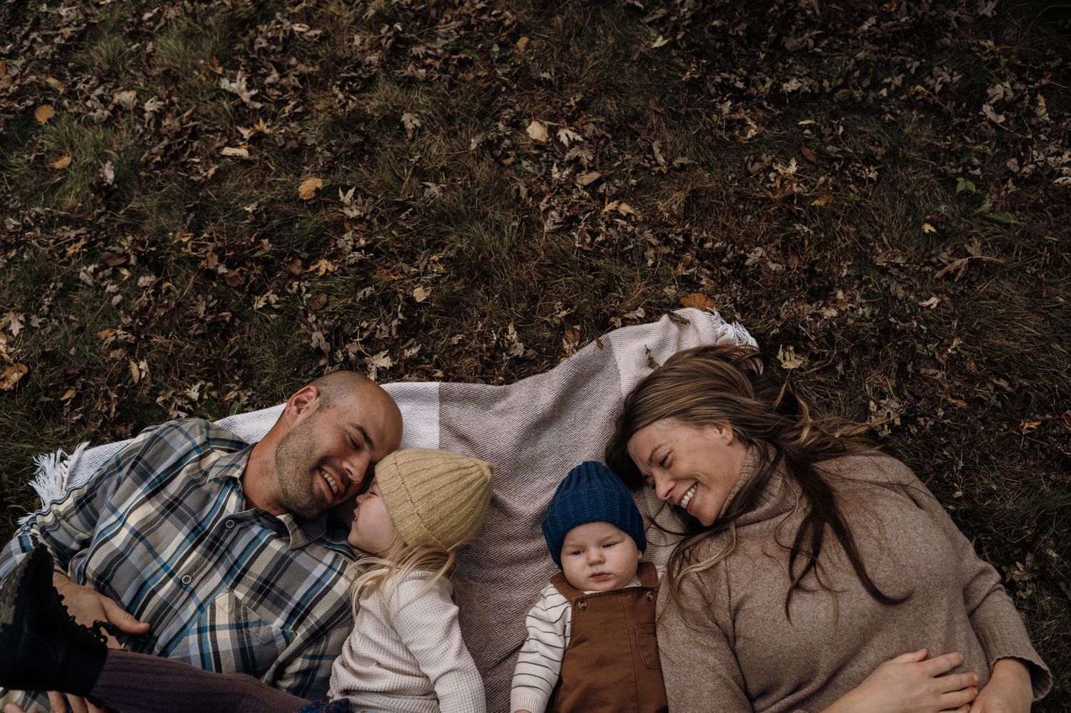 Family of four lies on a blanket covered in autumn leaves, parents snuggling two young children. Woodbury, Minnesota.
