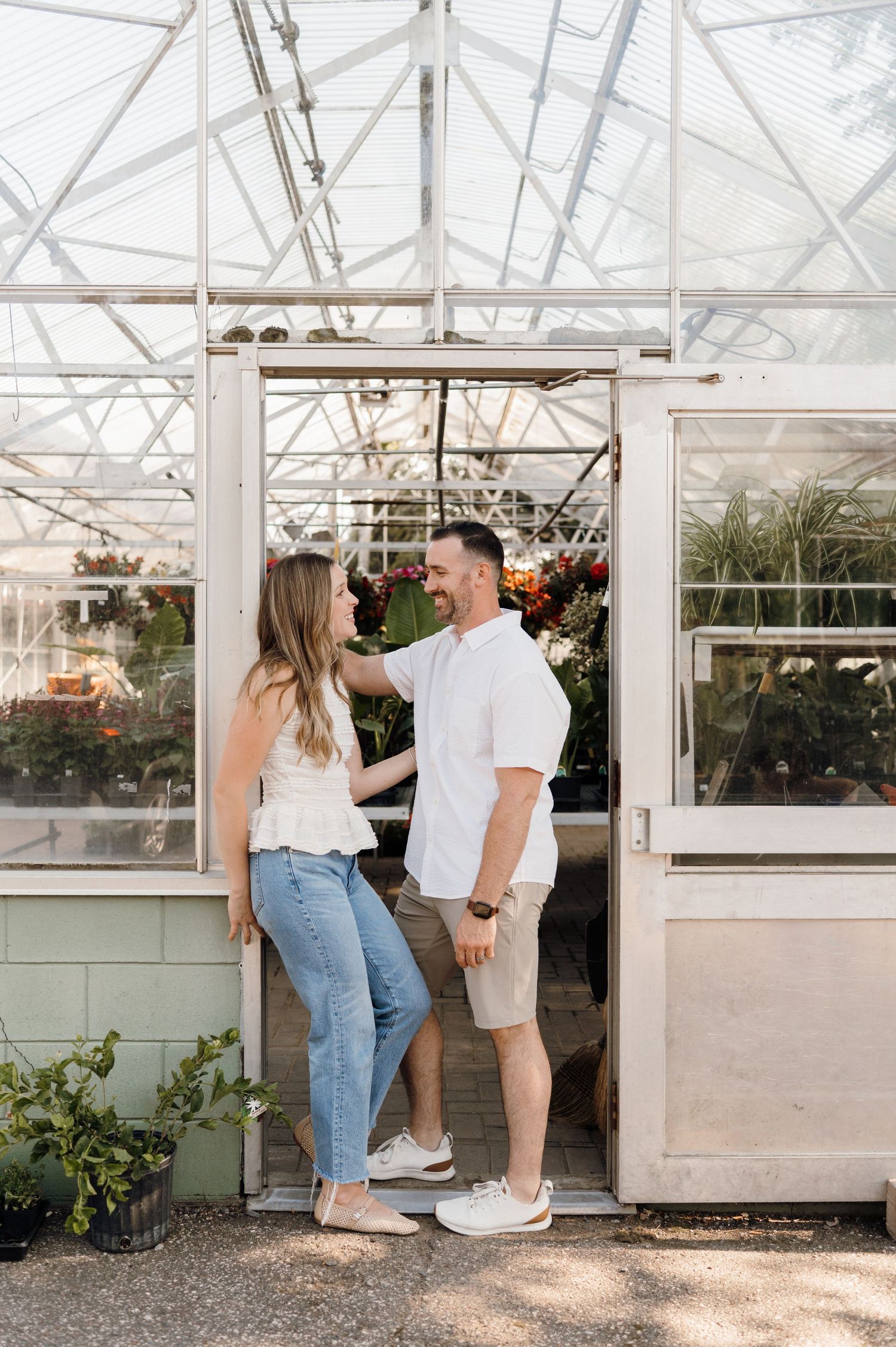 Smiling couple standing in greenhouse doorway looking at each other surrounded by plants.