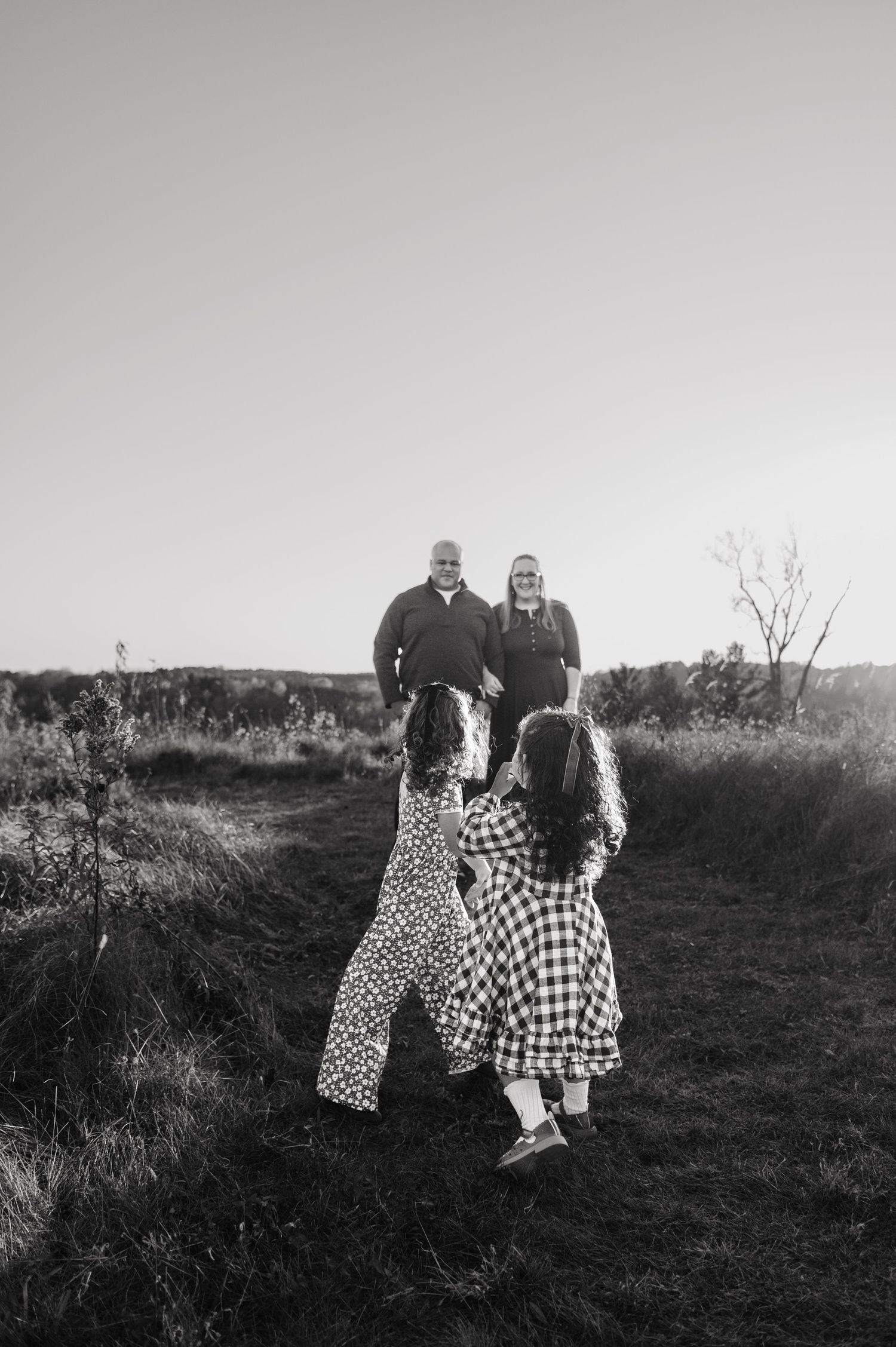 Black and white photo of two girls running toward parents on a grassy field path. Hudson, Wisconsin.