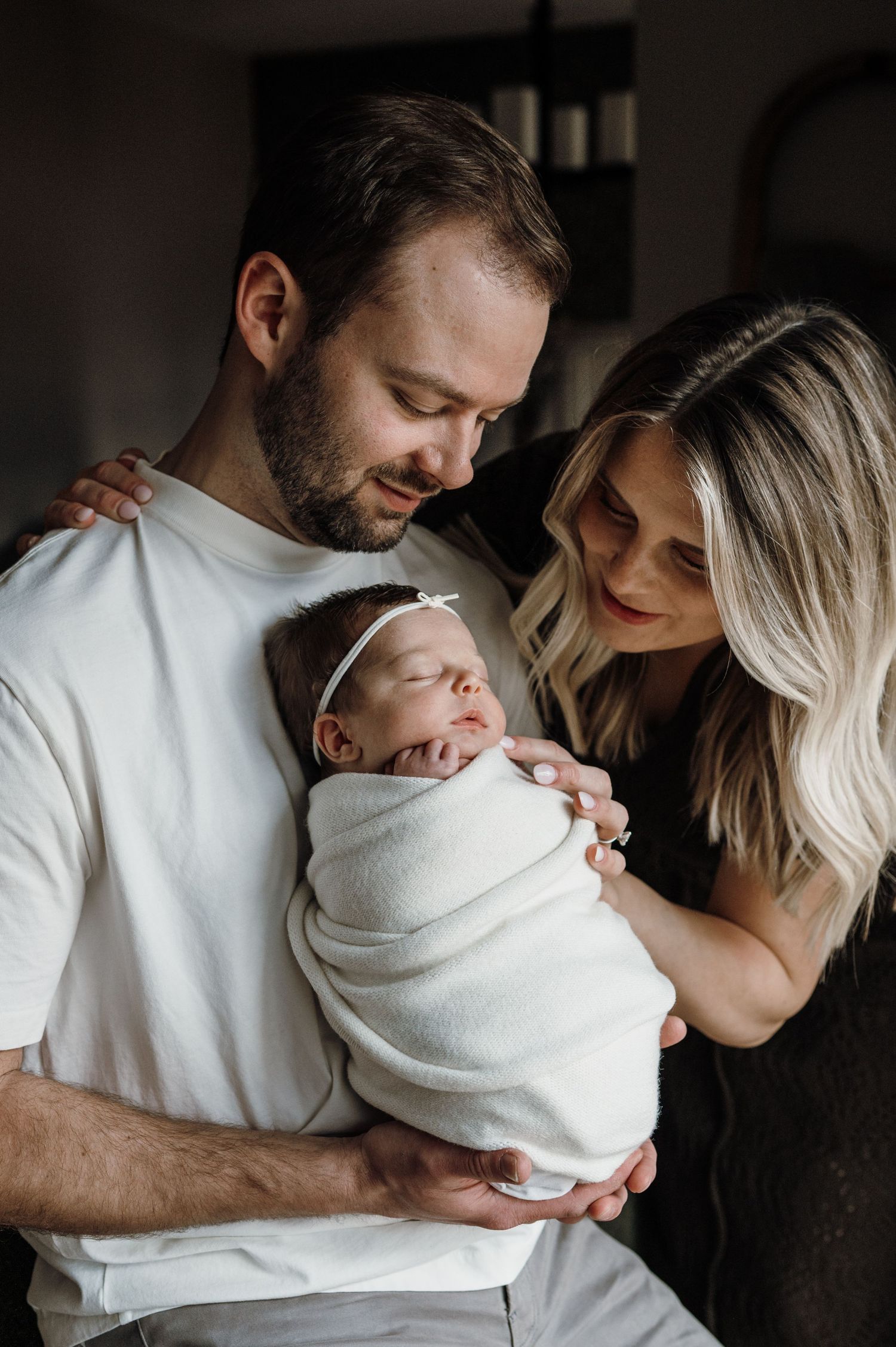 New parents holding swaddled newborn baby girl with white headband in cozy home setting.