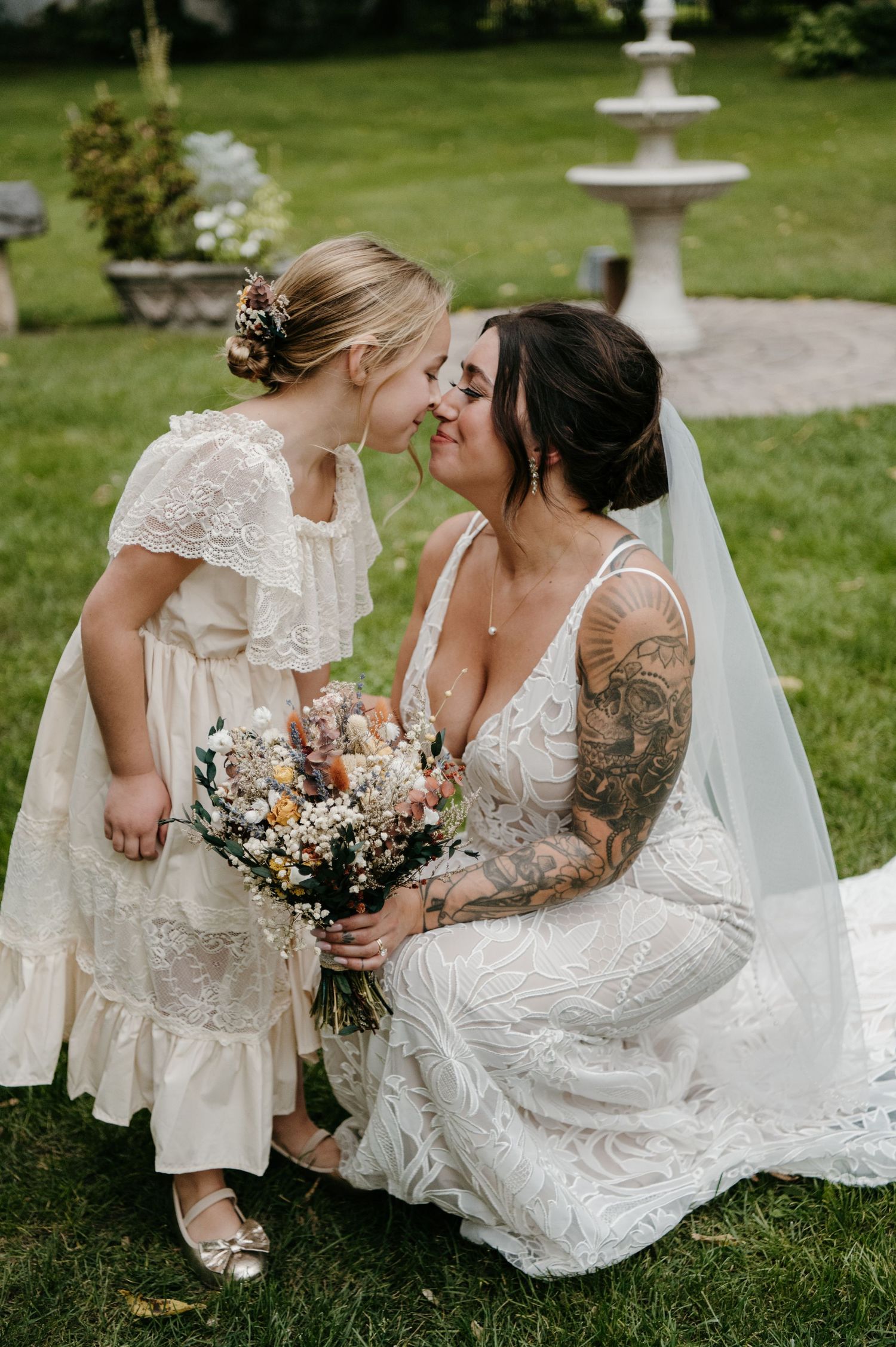 Tattooed bride in lace gown sharing sweet moment with flower girl outdoors. Wisconsin. Minnesota.