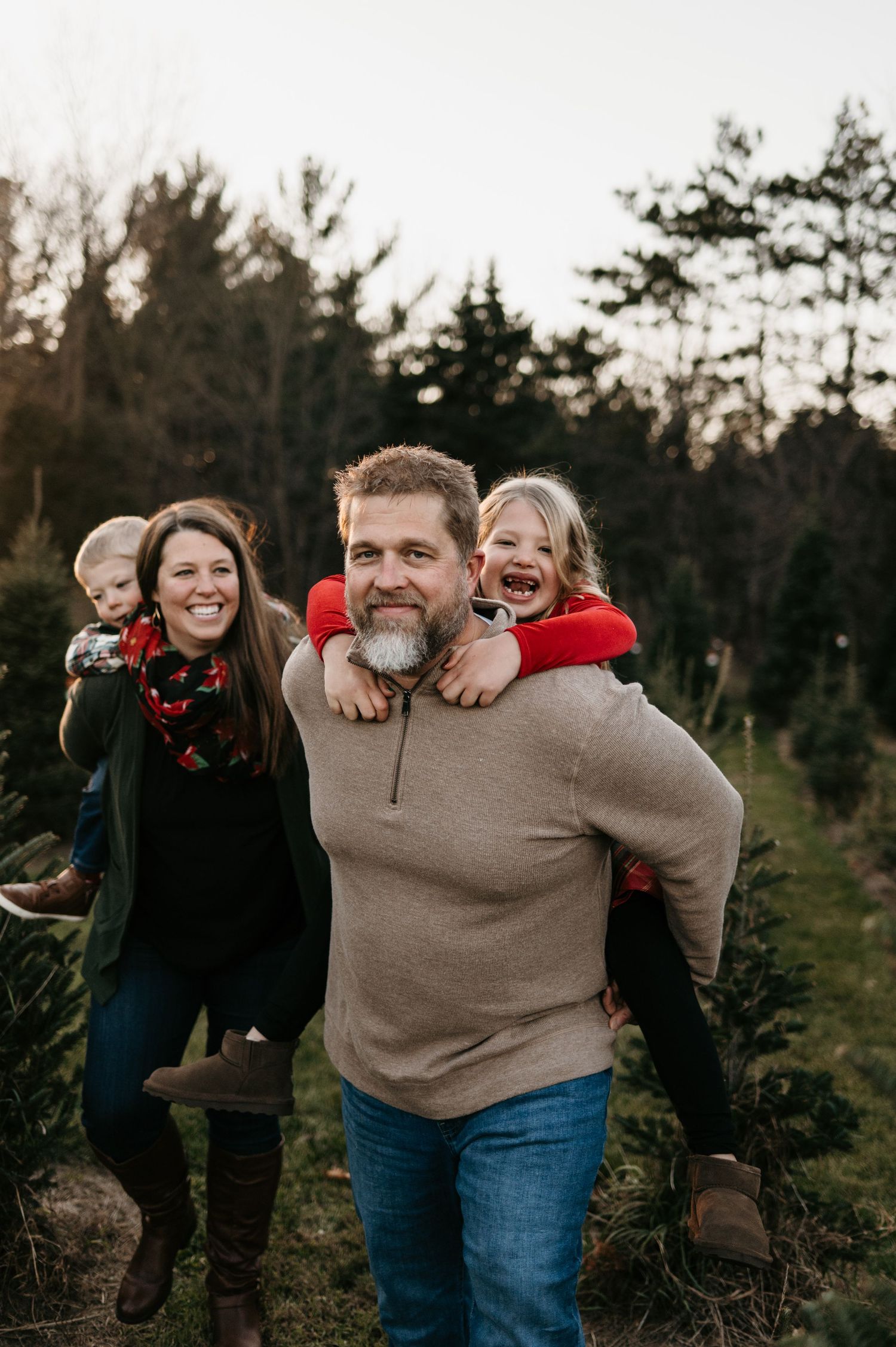 Joyful family at Christmas tree farm, dad giving daughter piggyback ride while mom holds baby. Hudson Wisconsin