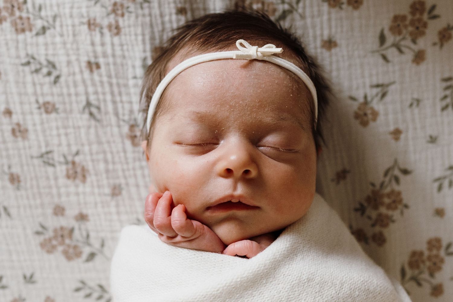 Sleeping newborn baby swaddled in white wrap with floral headband on floral fabric.