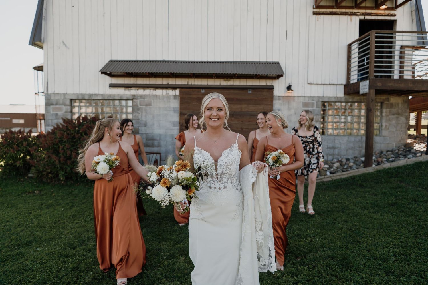 Smiling bride in lace gown leads bridesmaids in burnt orange dresses across green lawn at barn wedding venue.