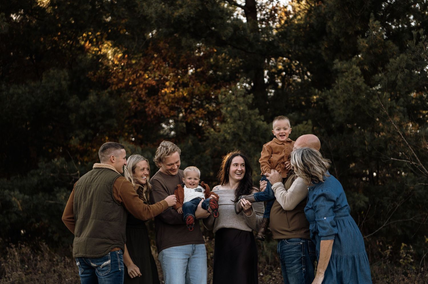 An extended family of seven adults and children gathers joyfully outdoors among autumn trees. Hudson, Wisconsin.