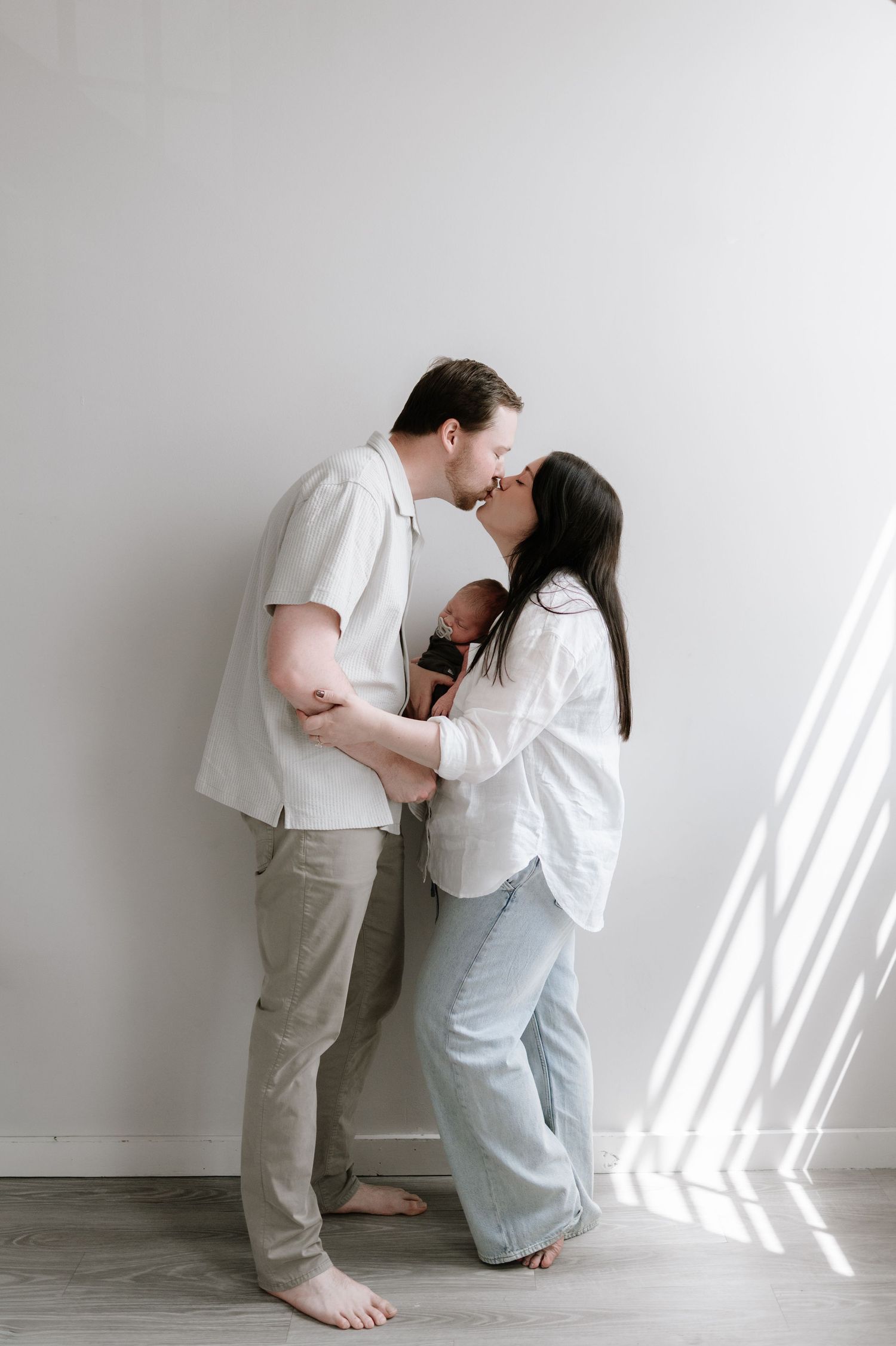 Couple kissing while holding their newborn baby in a bright minimalist white room.