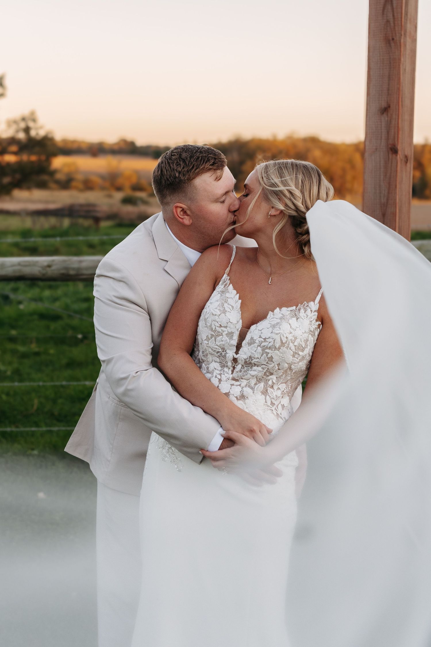 Groom in tan suit kisses bride in lace gown as veil flows in wind at rustic outdoor wedding. Wisconsin. Minnesota.
