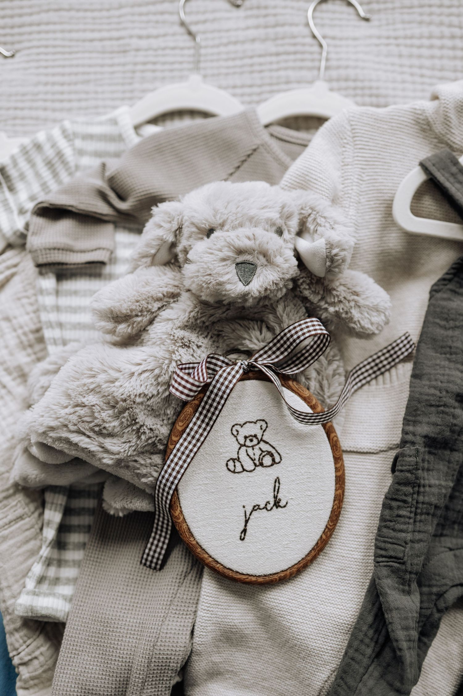 Baby name embroidery hoop with teddy bear beside neutral toned newborn clothes.