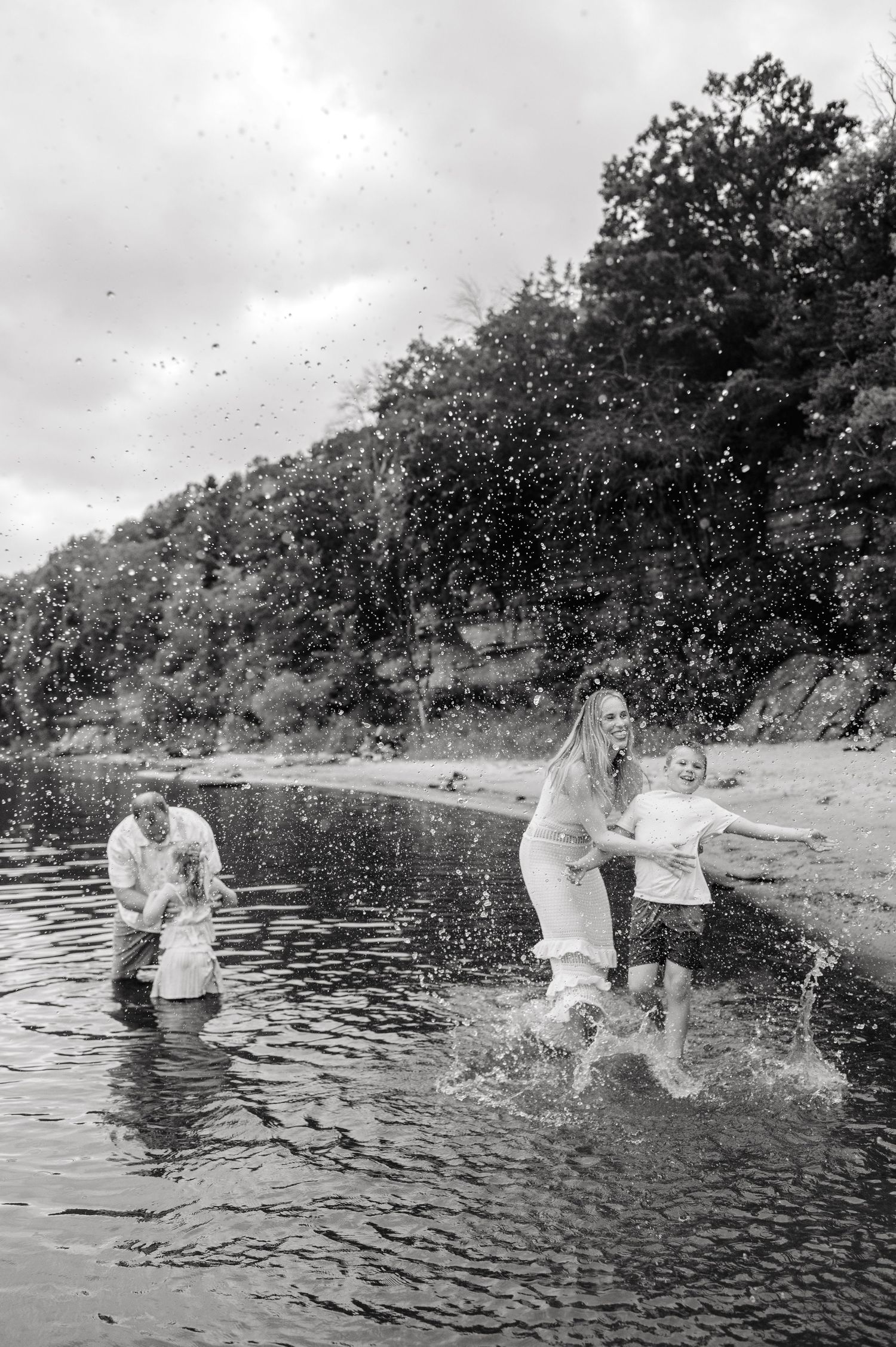 A joyful family splashes in a river in a black and white outdoor photo.