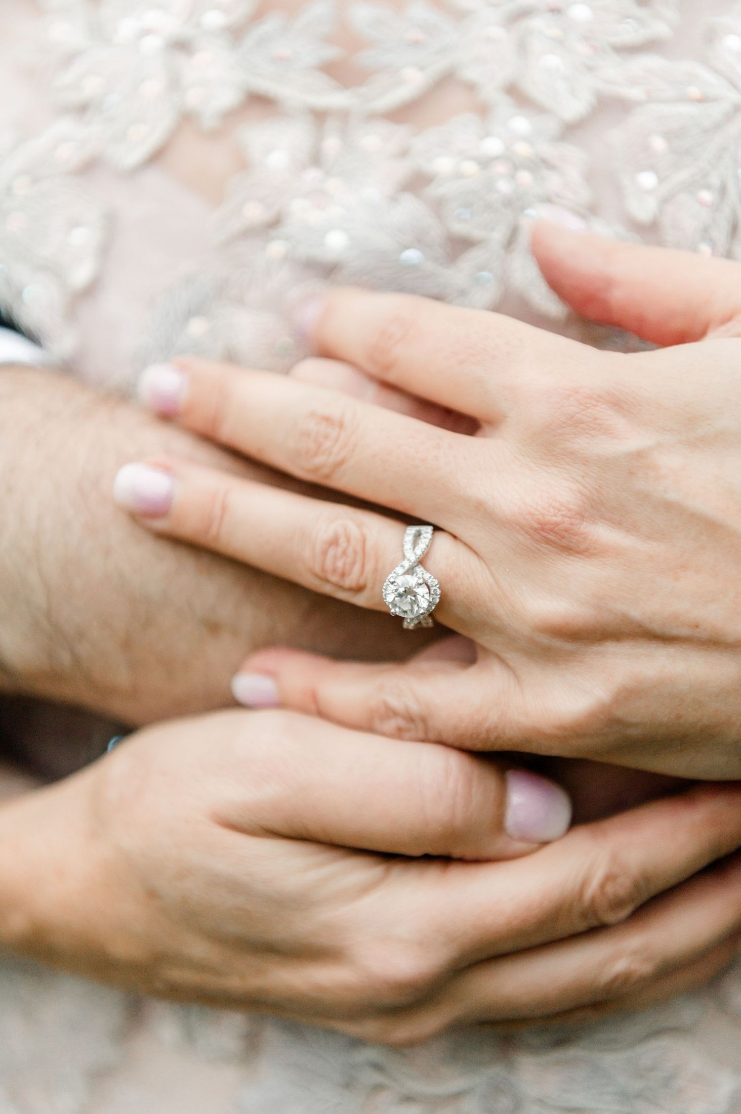 Close-up of diamond engagement ring on bride's hand over lace wedding dress.