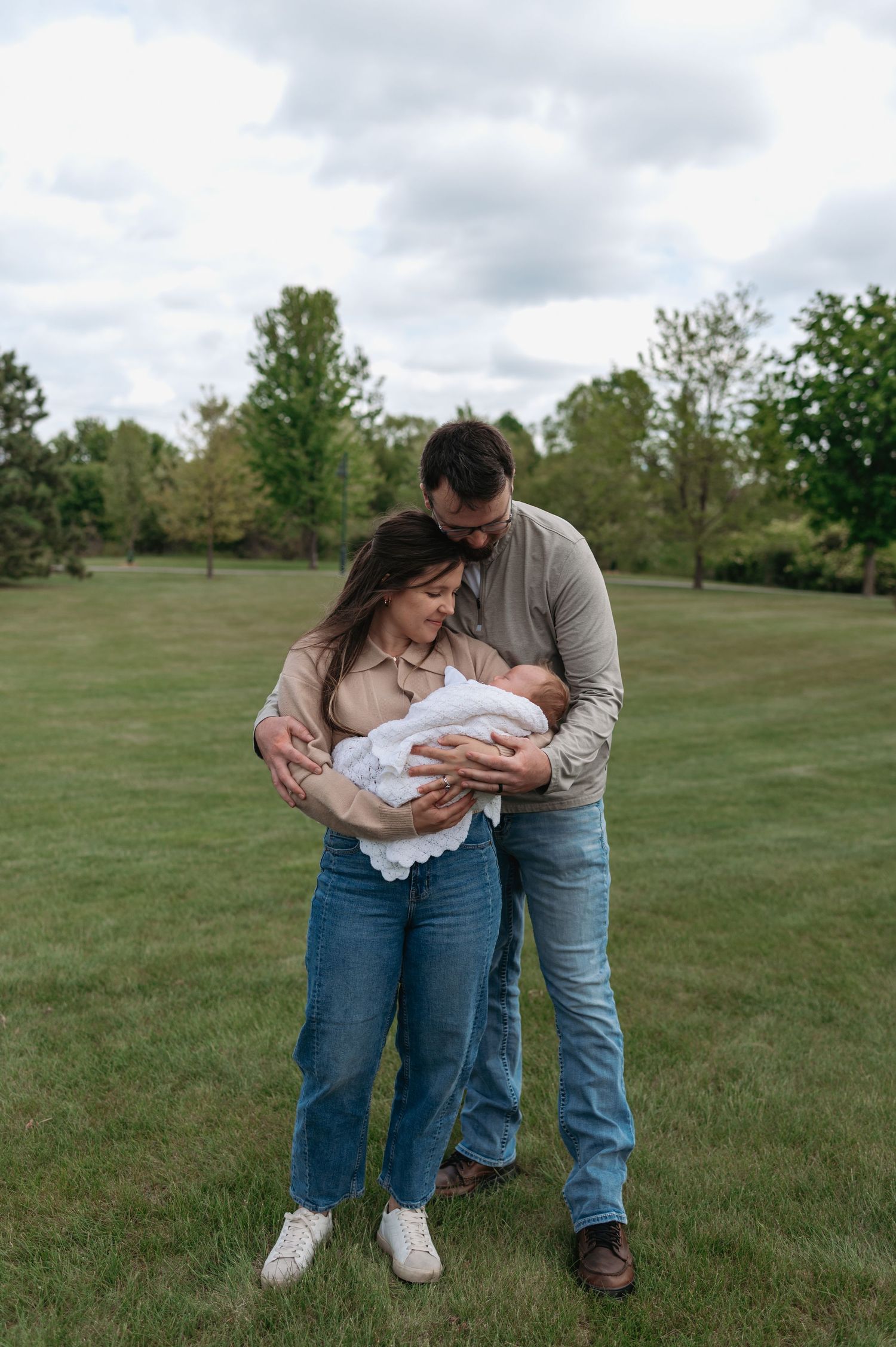 Young couple standing in green park cradling newborn baby wrapped in white blanket on cloudy day.
