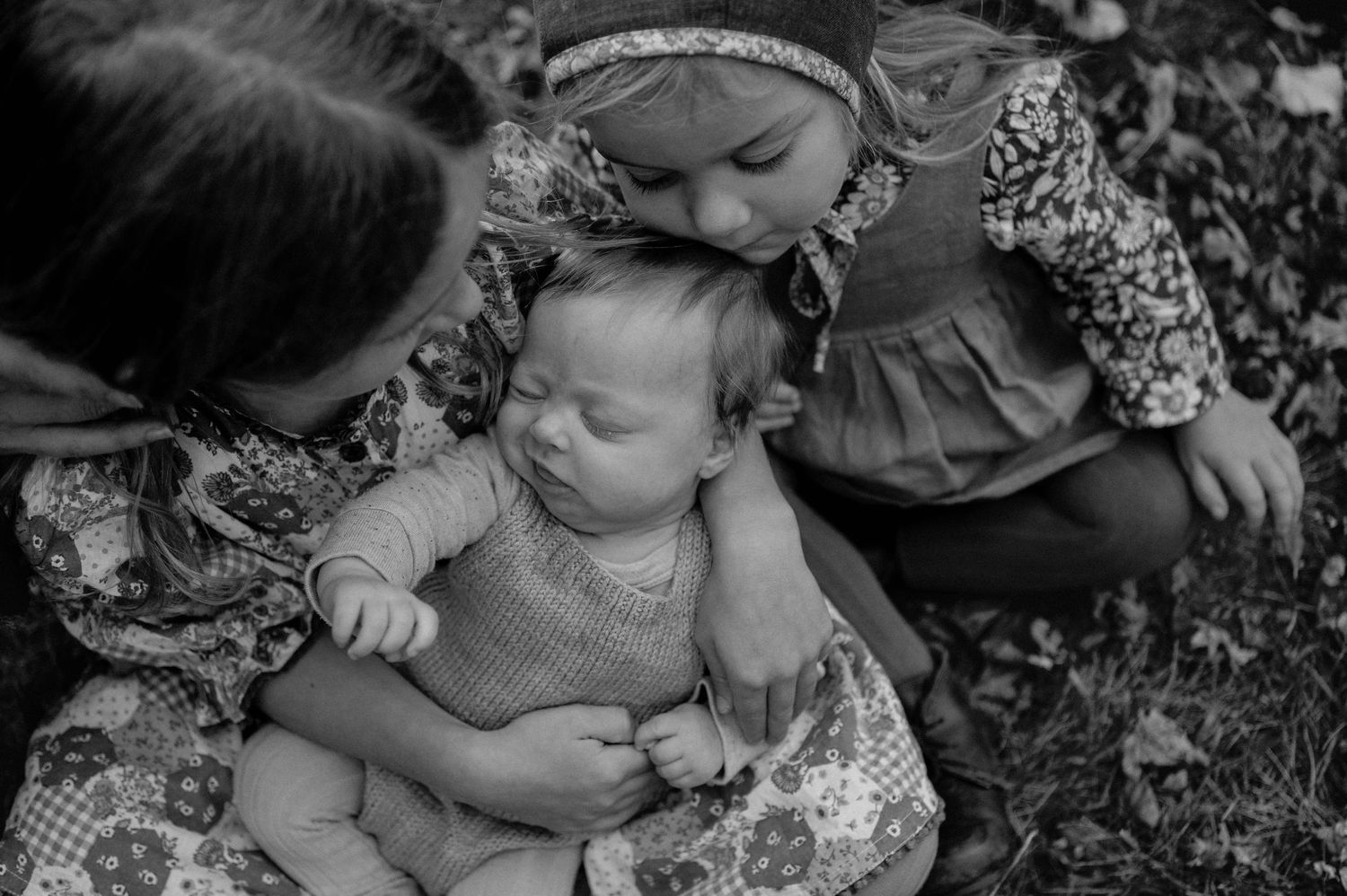 Two girls tenderly kiss and hold a sleeping newborn baby outdoors in a black and white family photo. Woodbury, Minnesota.