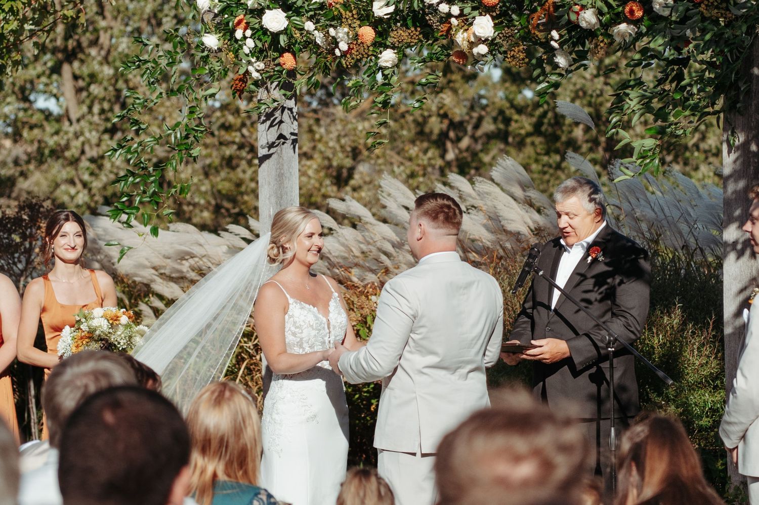Bride and groom exchange vows beneath floral arch at outdoor wedding ceremony with guests seated. Wisconsin. Minnesota.