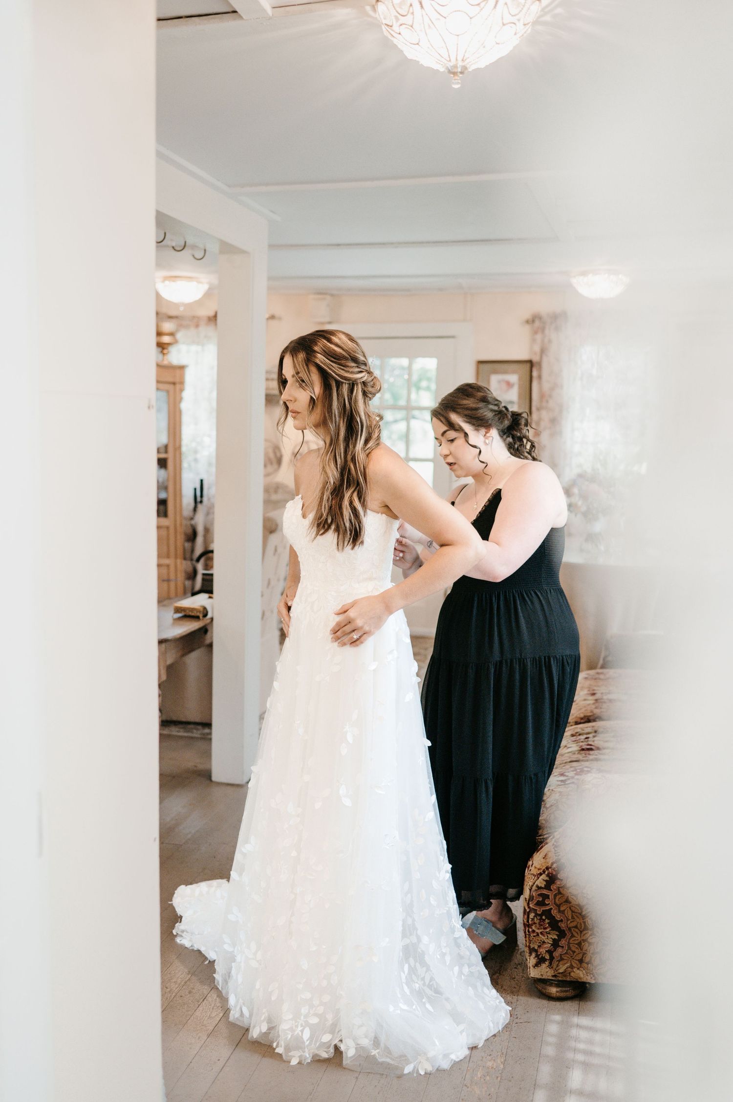 Bridesmaid in black dress helping bride into floral applique wedding gown in bright bridal suite.