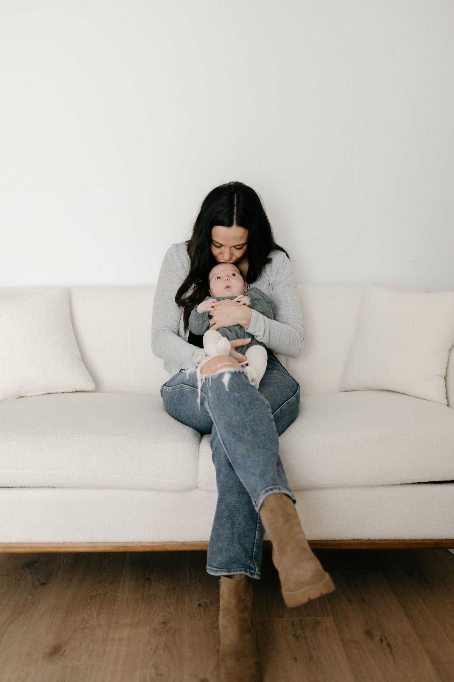 Dark-haired mother in gray top and jeans tenderly kissing her newborn baby on a white sofa.