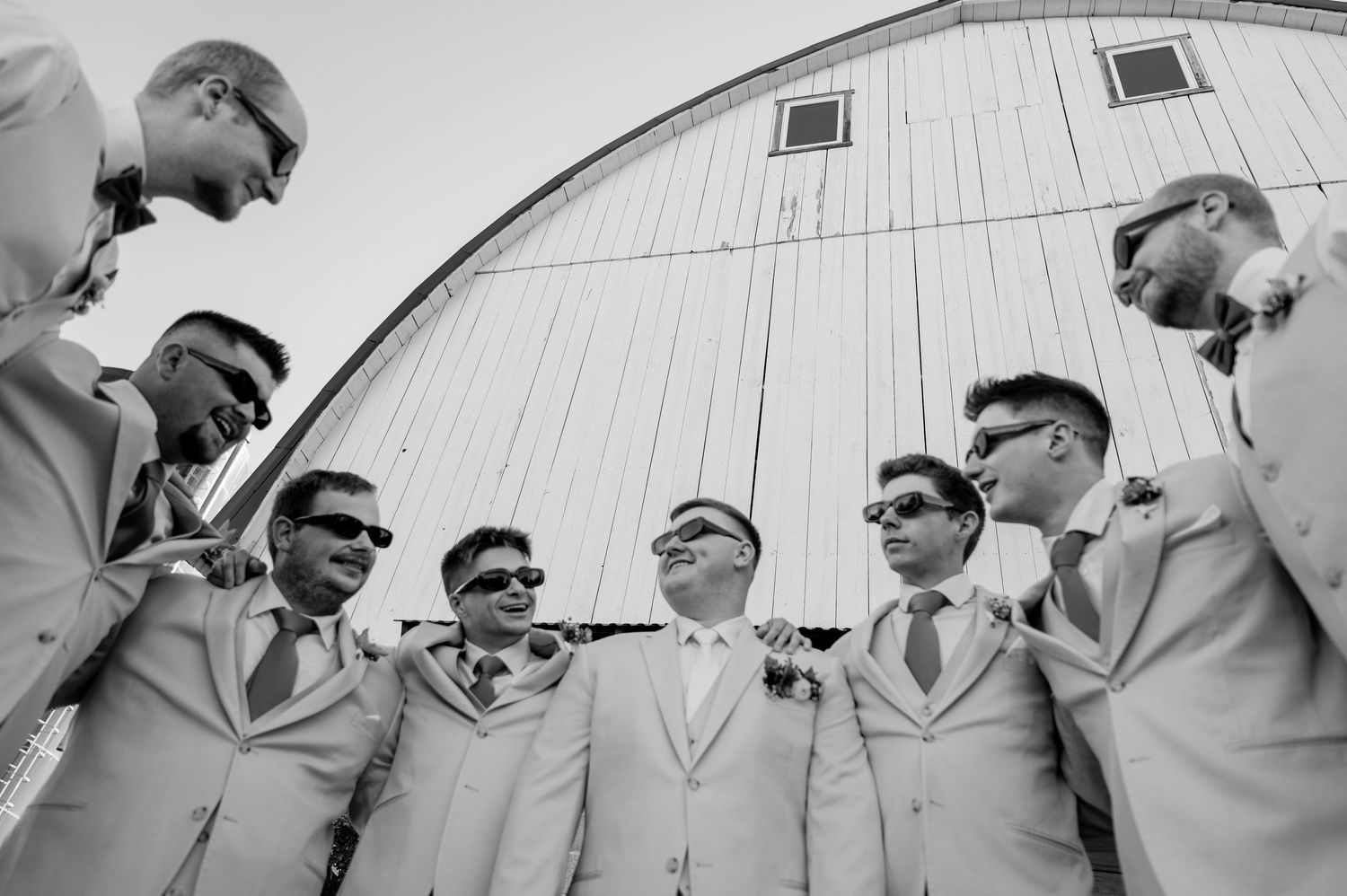 Black and white photo of groom and groomsmen in light suits and sunglasses gathered in front of white arched barn.